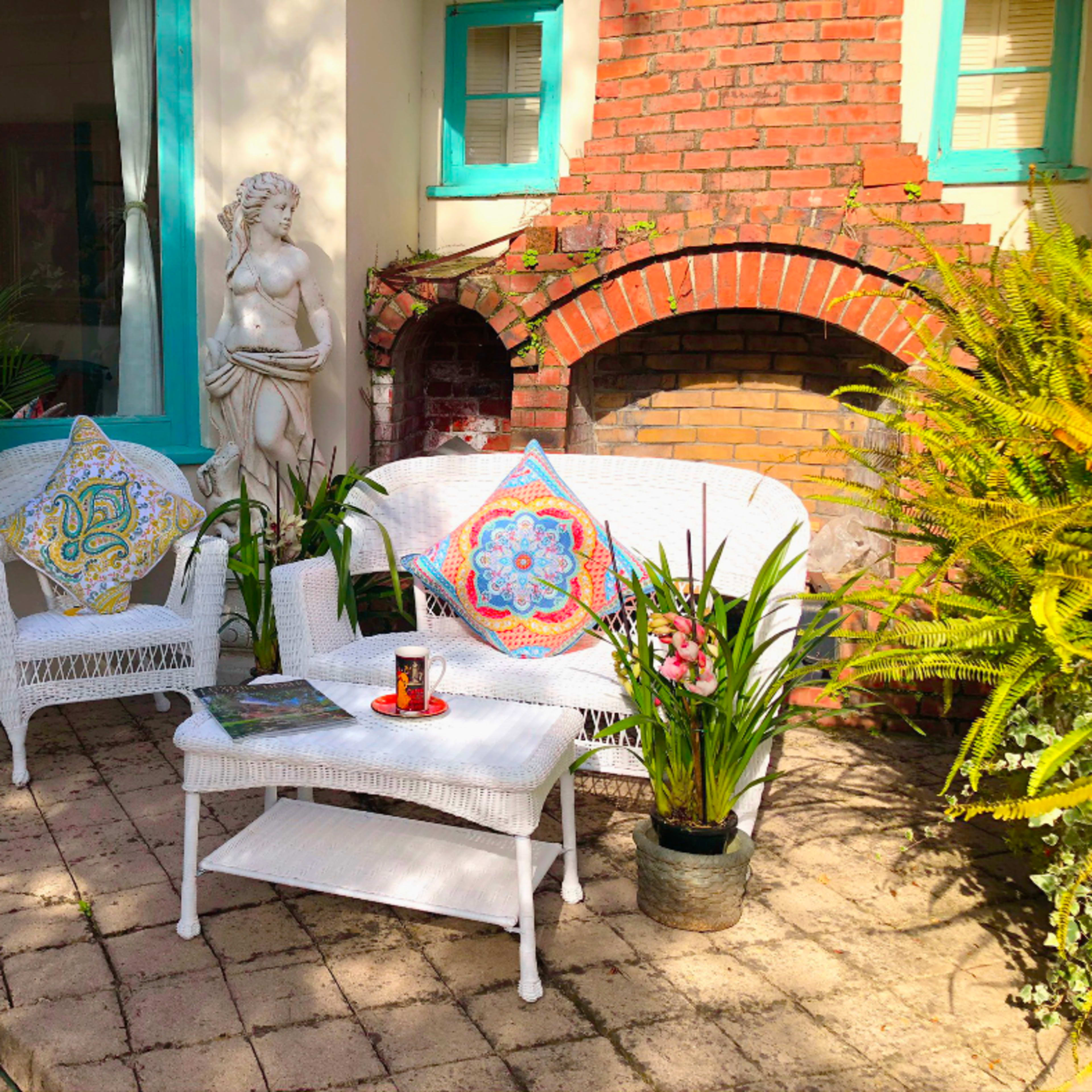 A cozy outdoor seating area features white wicker furniture, colorful cushions, a small table with a candle, and greenery around a brick wall with a sculpture.