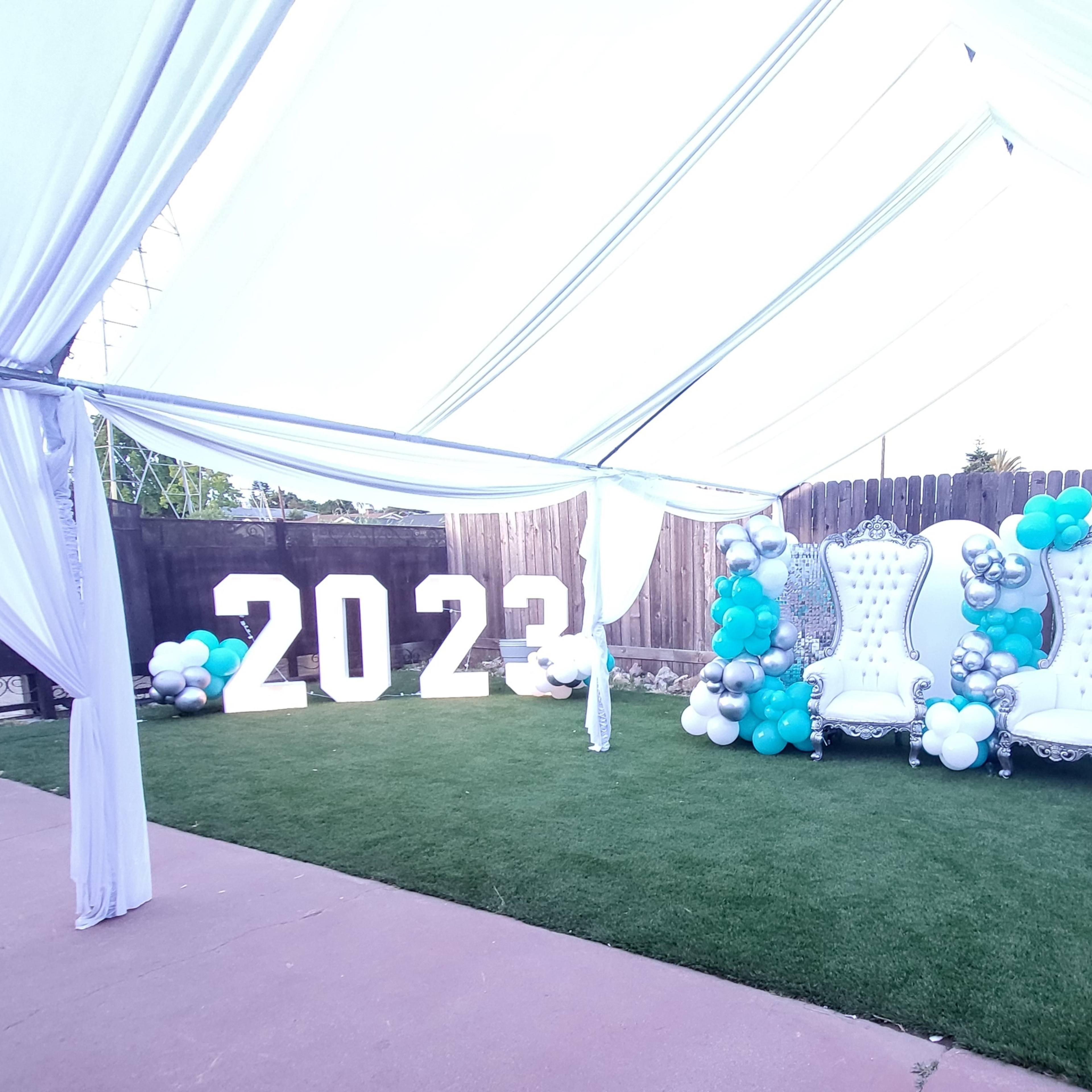 A decorated area for a prom event featuring large "2023" numbers, two white throne chairs, and an arrangement of balloons in teal, silver, and white.