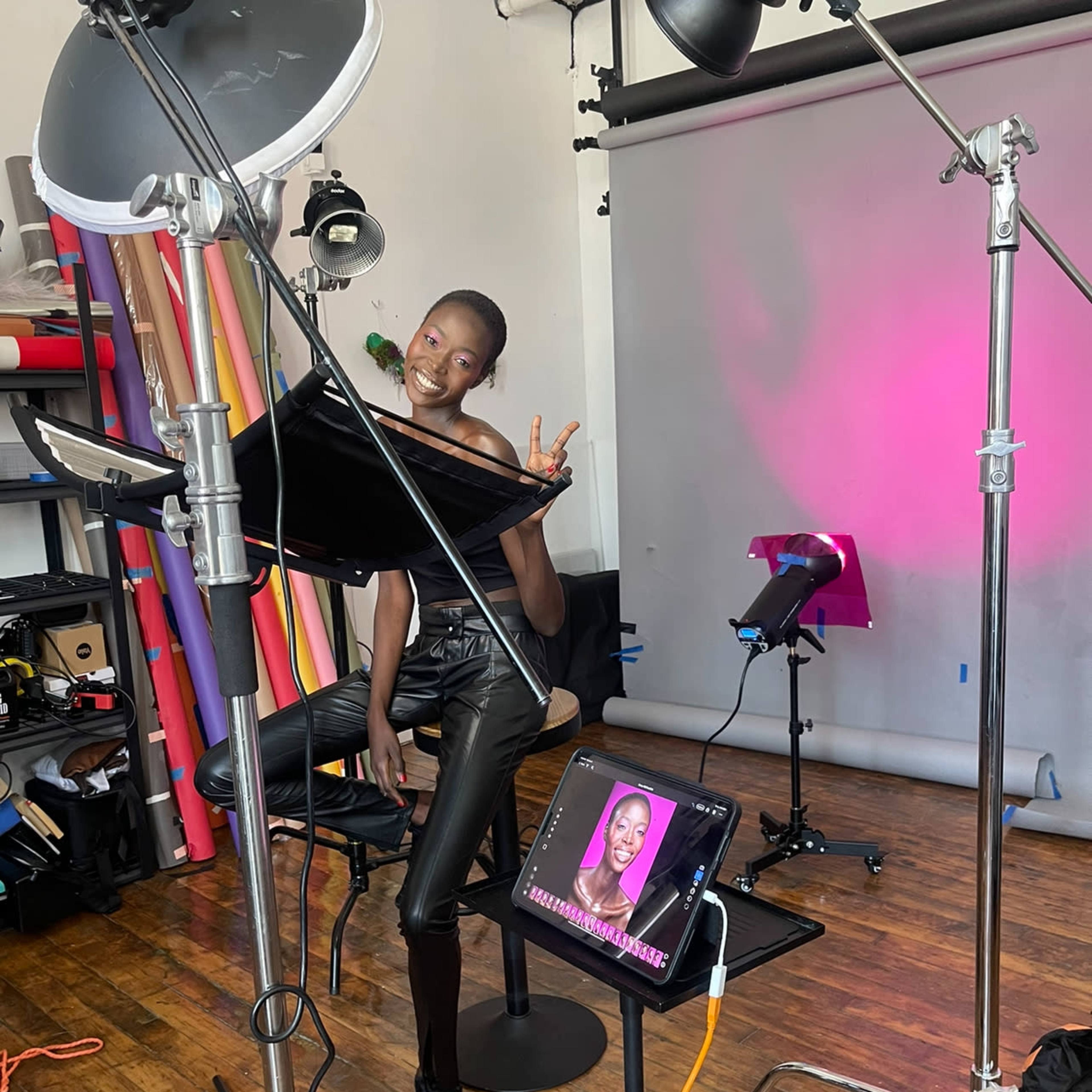 A model strikes a pose in a photography studio surrounded by lighting equipment and backdrop rolls.