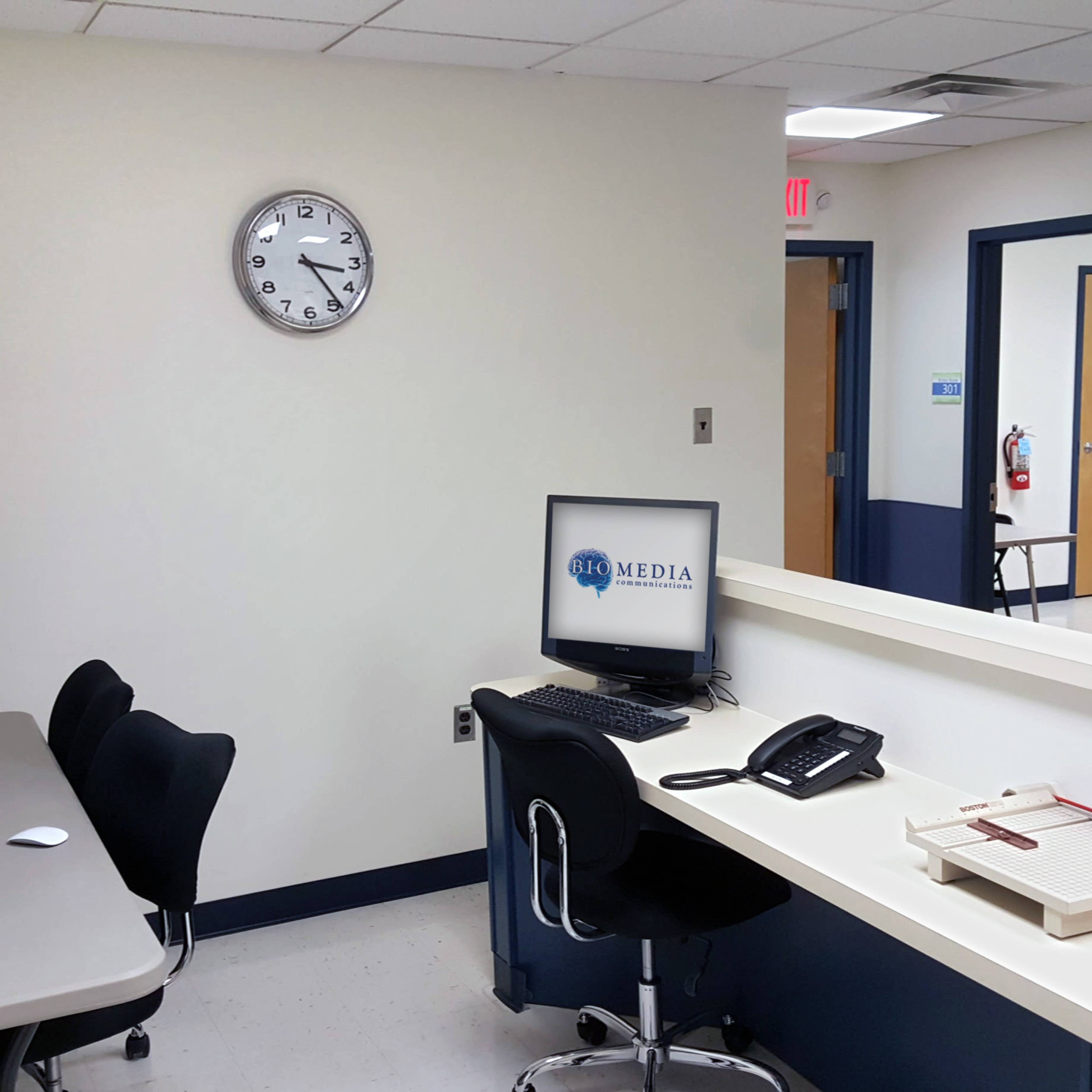 A neatly arranged office space featuring a wall clock, computer stations, a telephone, and a receptionist desk with a wooden registration board.