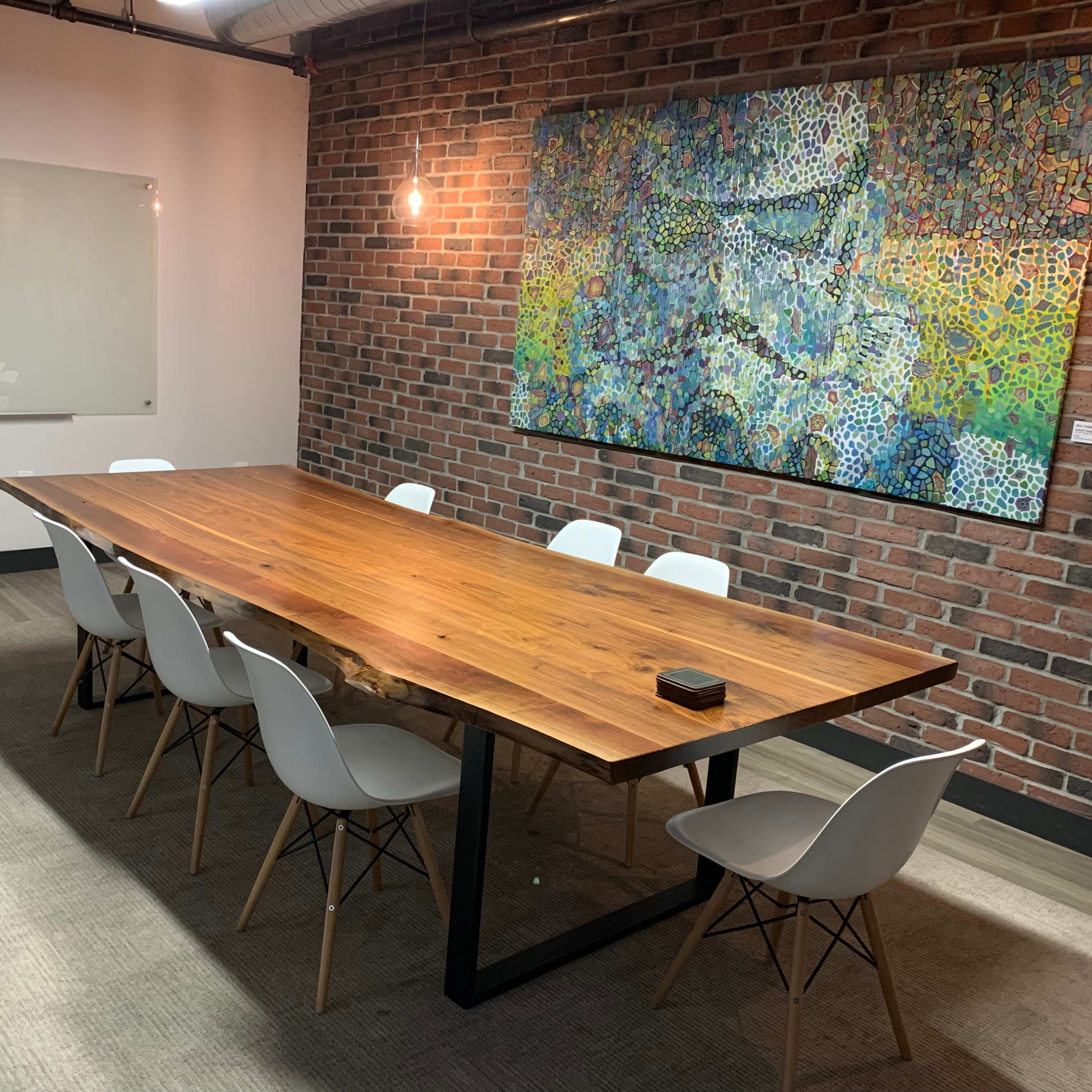 A conference room features a long wooden table surrounded by white chairs, with a colorful artwork displayed on a brick wall.