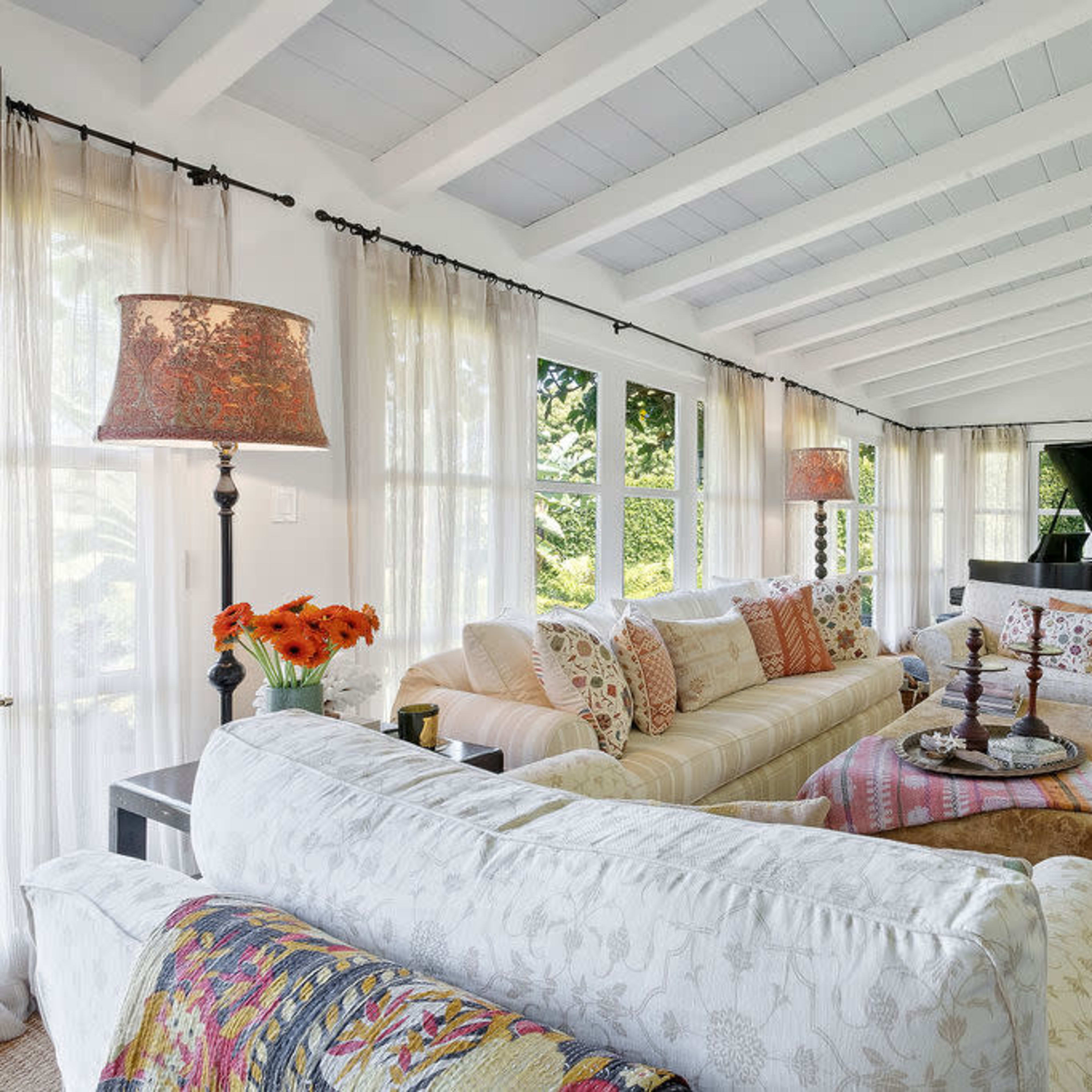 A sunlit living room features white-beamed ceilings, large windows with sheer curtains, and a grand piano in the background, surrounded by comfortable seating and a decorative rug.
