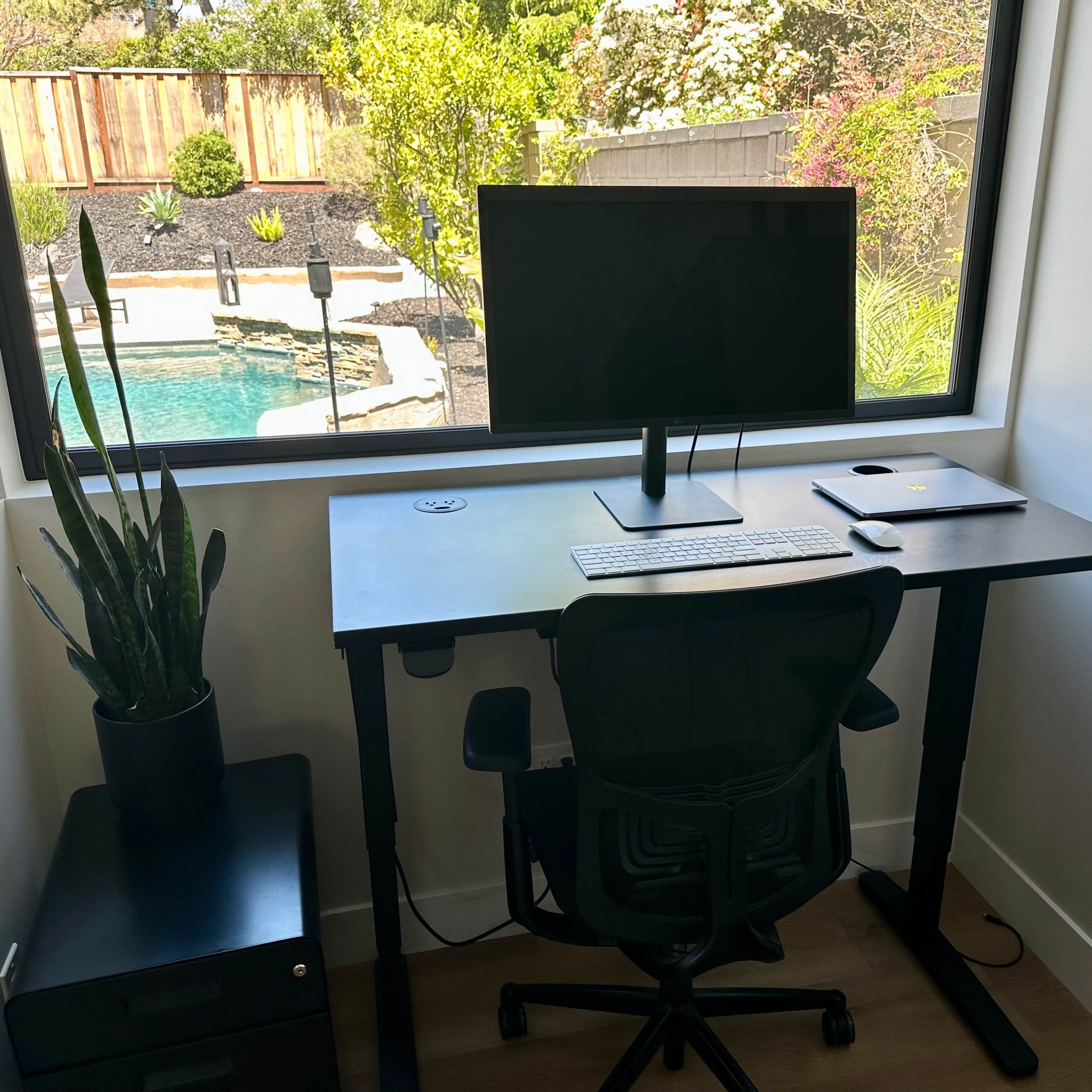 A modern workspace is set up beside a large window, featuring a desk with a computer monitor, keyboard, and laptop, while a tall plant and a small cabinet are nearby.