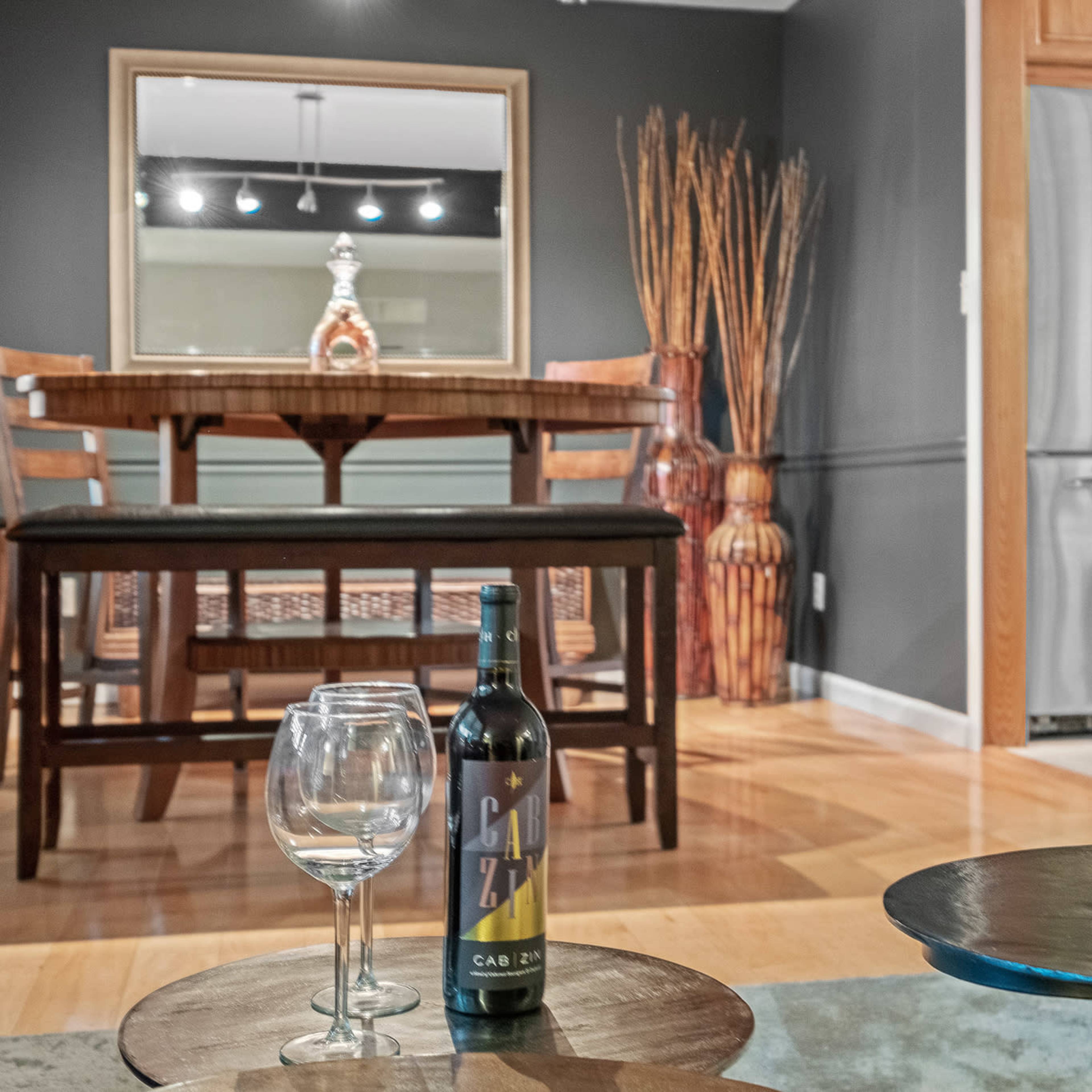 A modern dining area featuring a wooden table with chairs, a wine bottle and glasses on a foreground table, and decorative elements like a vase and an ornate mirror on the wall.