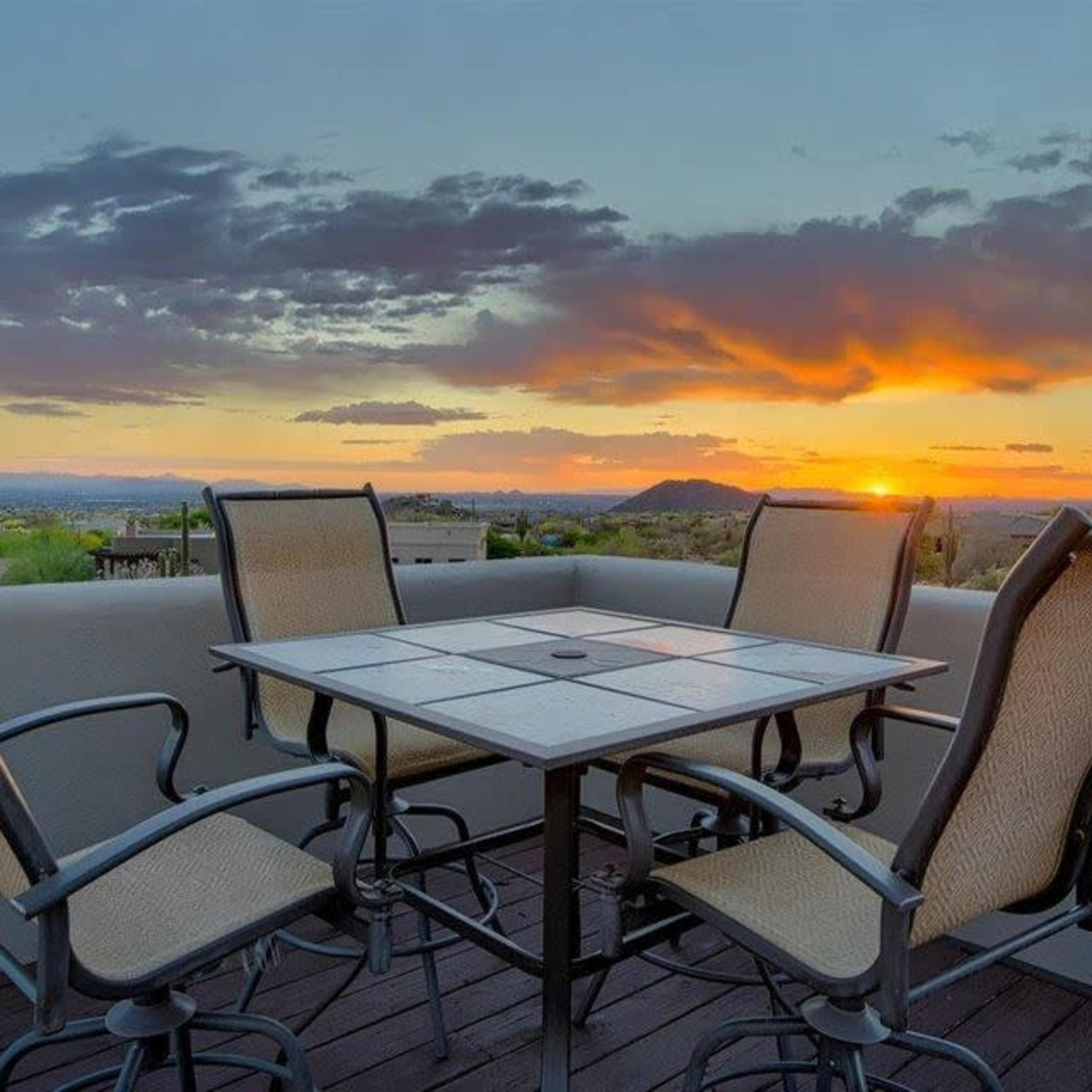 A patio set with four chairs surrounds a square table, overlooking a sunset with mountains and clouds in the background.