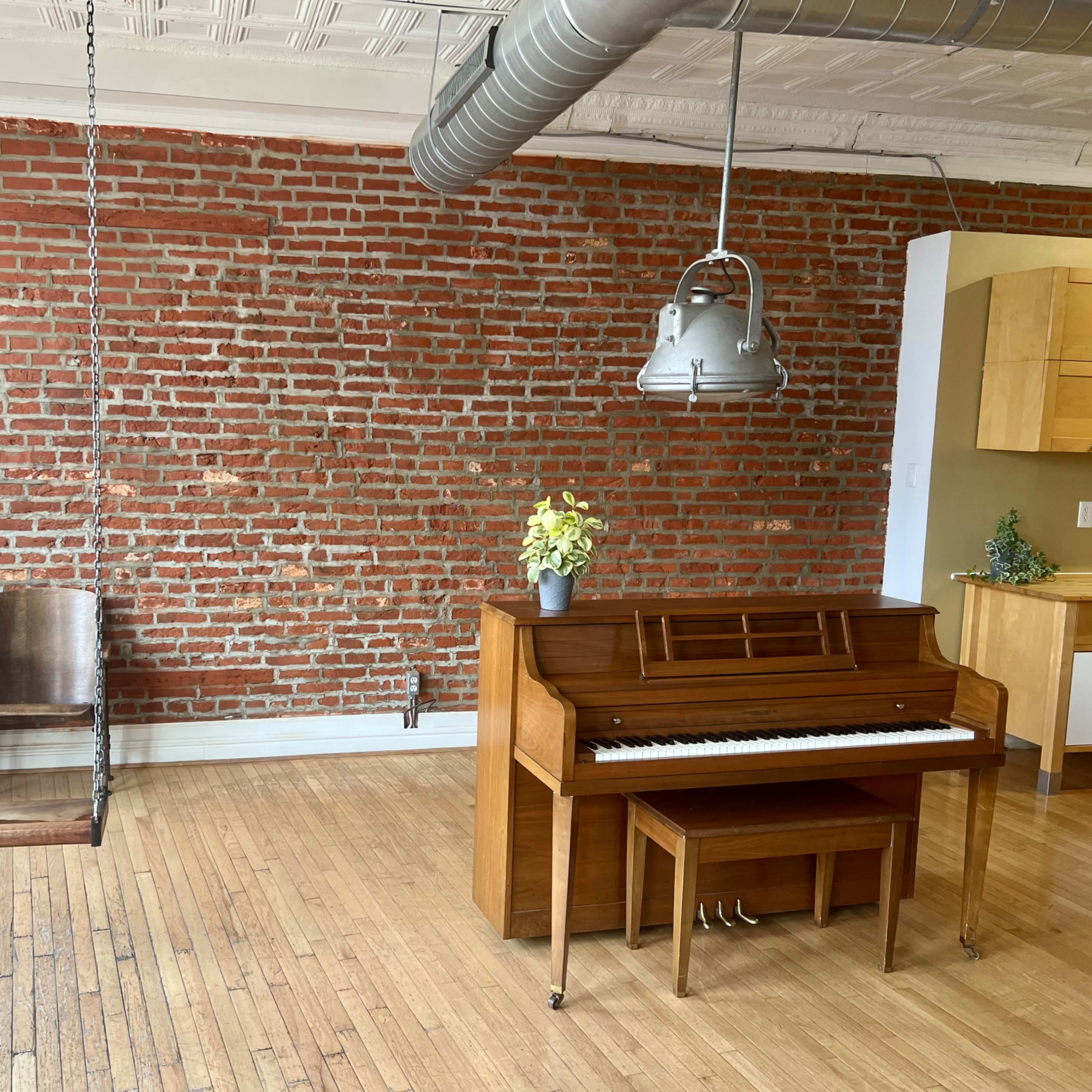 A wooden piano and a hanging swing chair are placed against a brick wall in a spacious room with wooden flooring.
