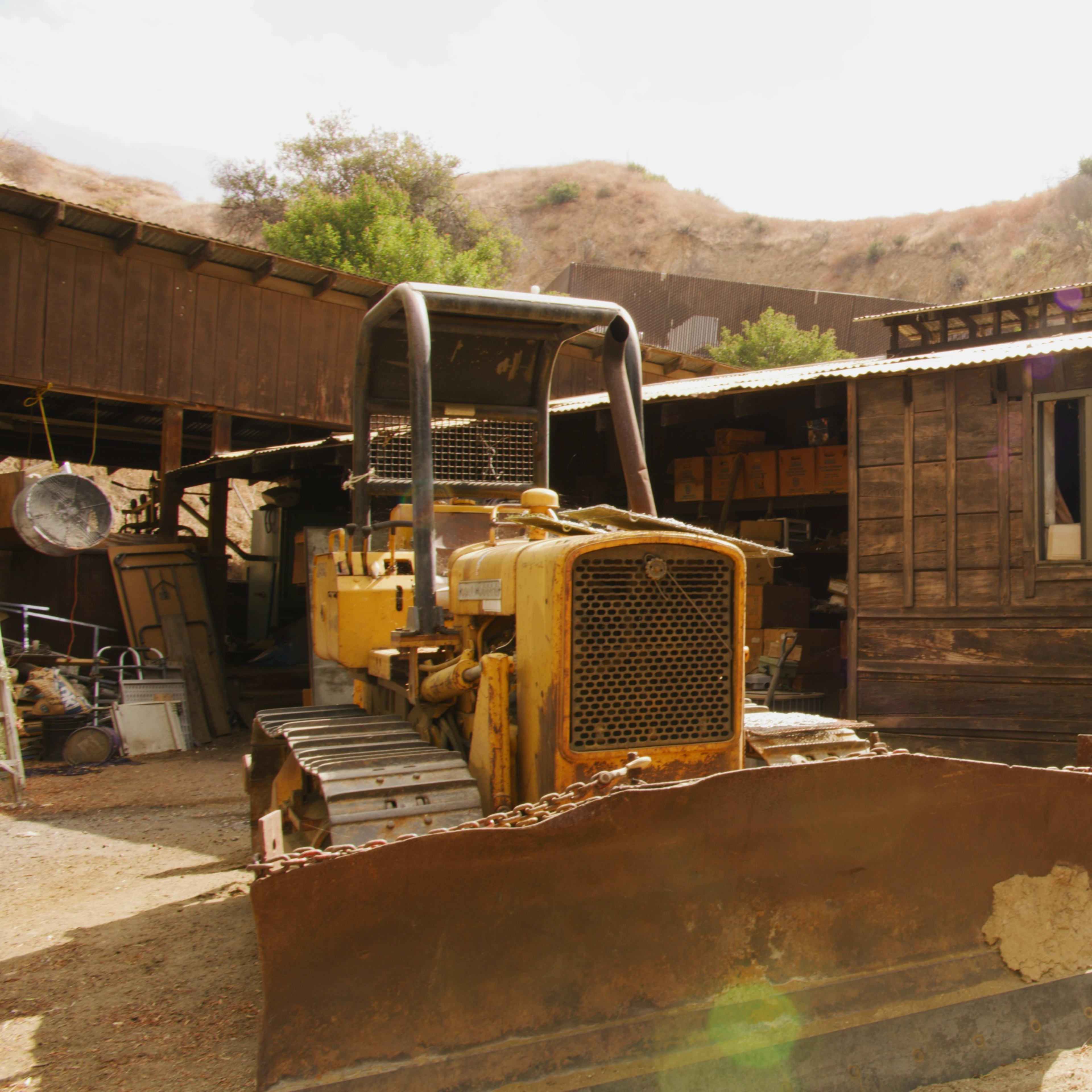 A yellow bulldozer is parked in front of a wooden shed with a mountain backdrop and several items scattered around the yard.