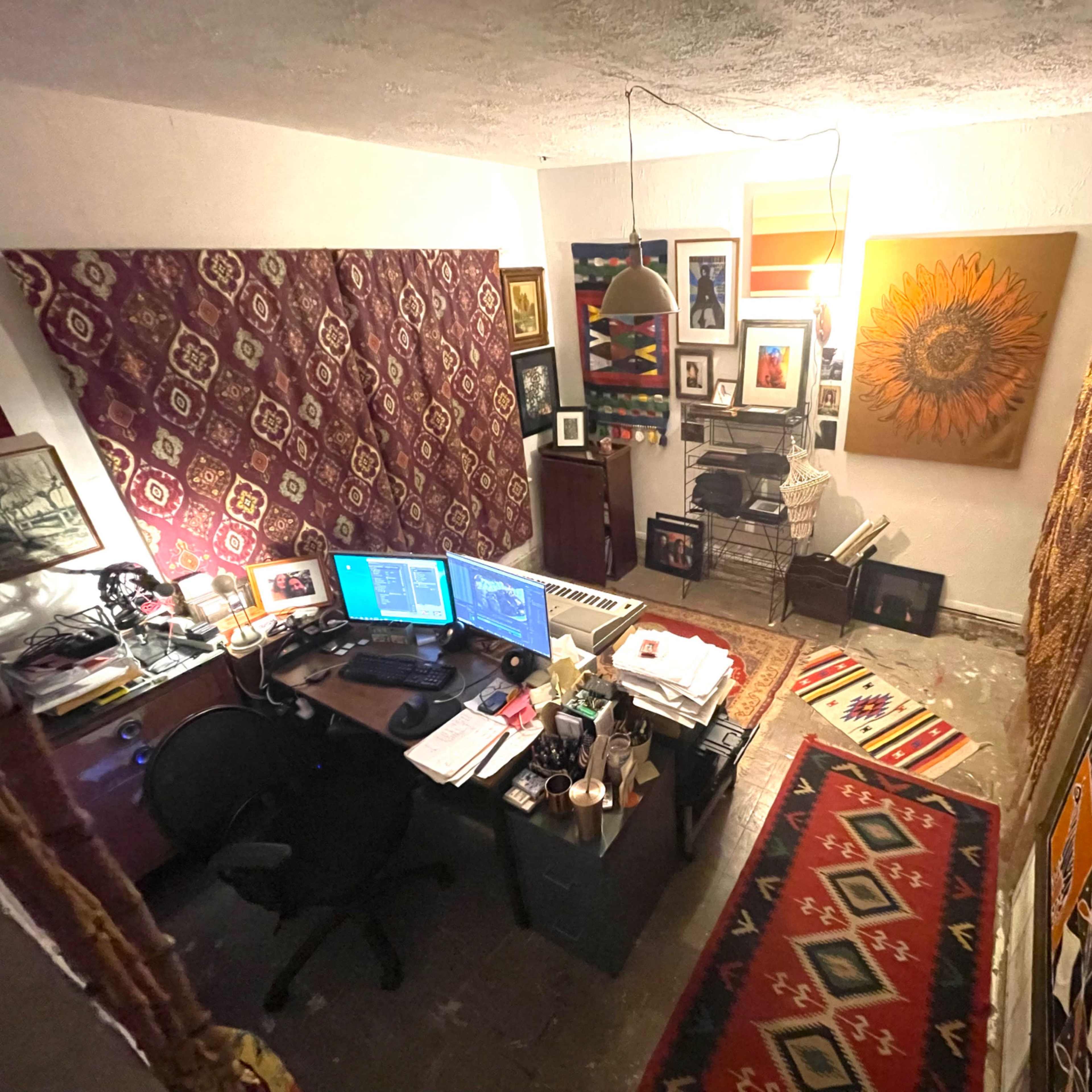 A small home office features a desk with two computer monitors, surrounded by colorful wall hangings and decorative art.