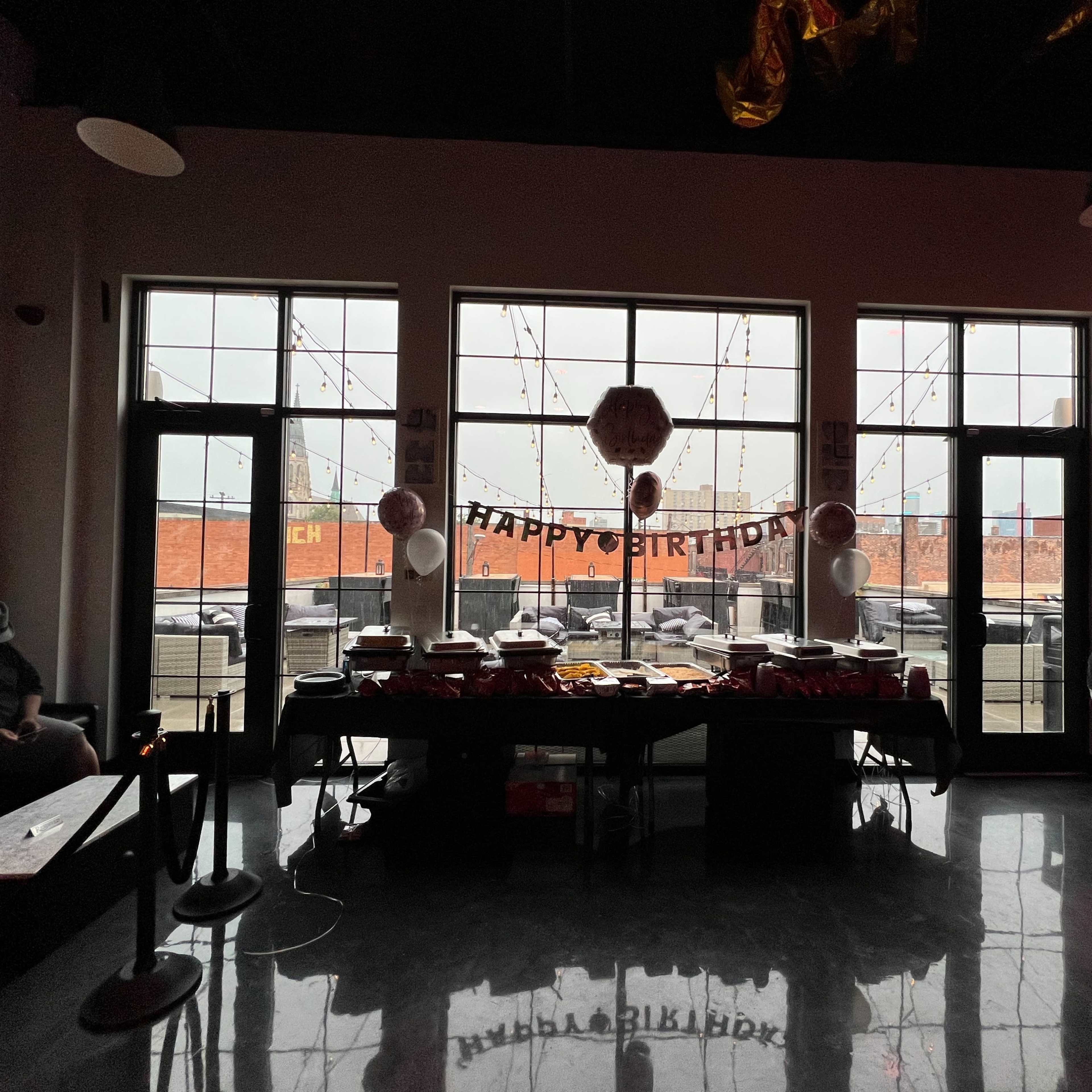 A birthday celebration setup features a table with food, decorations, and a "Happy Birthday" banner, with large windows overlooking an outdoor area.