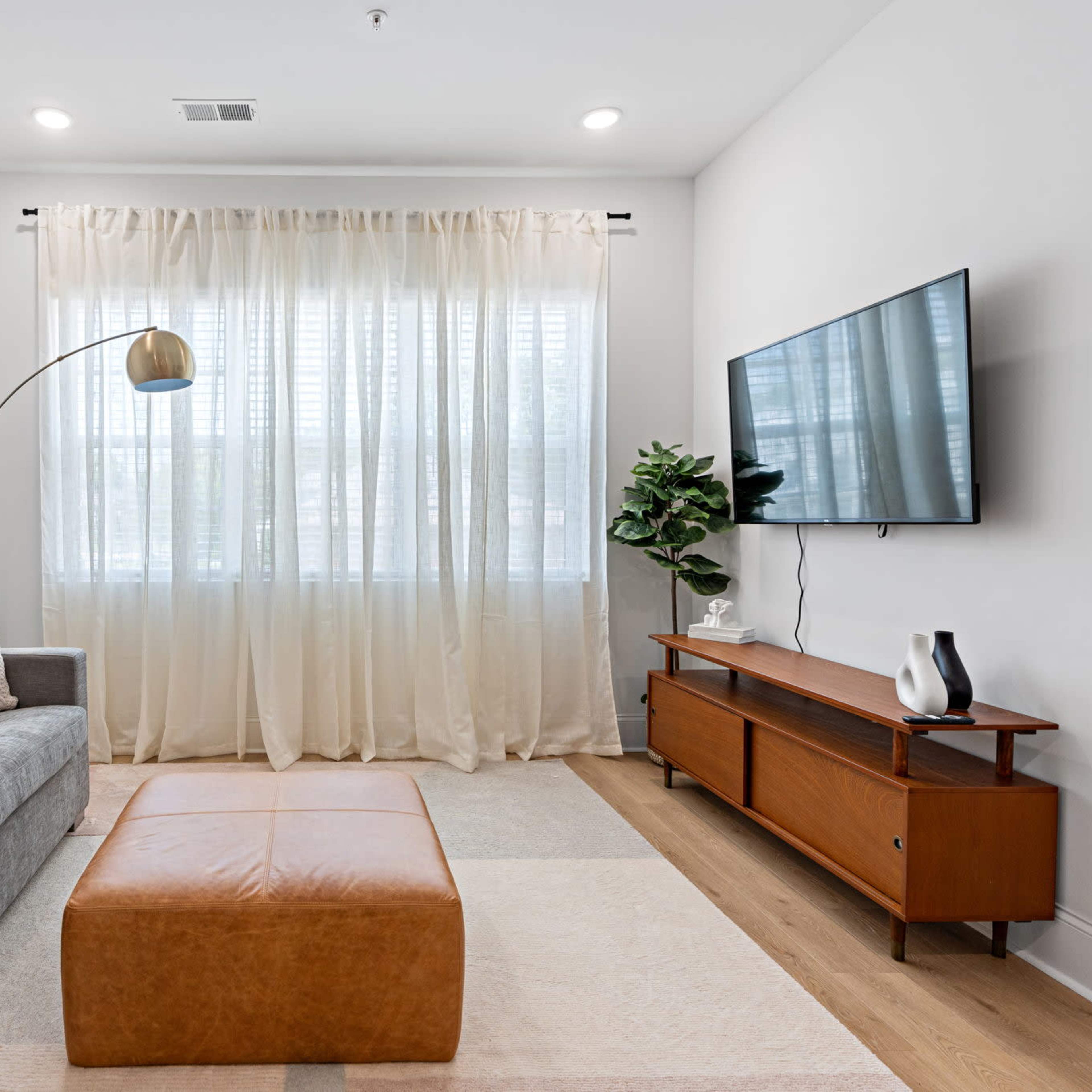 A modern living room featuring a gray sofa, a brown leather ottoman, a wooden console table, a large television mounted on the wall, and sheer curtains allowing natural light to enter.