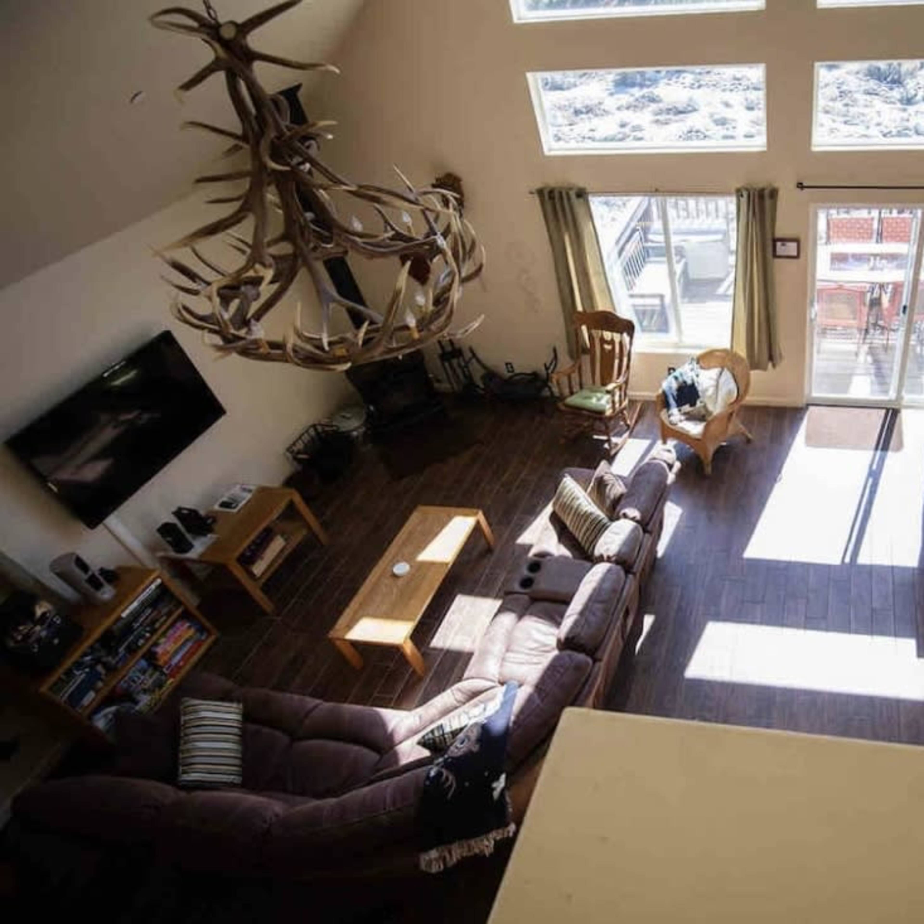 A spacious living area with high ceilings, featuring a large window, a dining area with wooden tables and chairs, and a seating arrangement centered around a television.