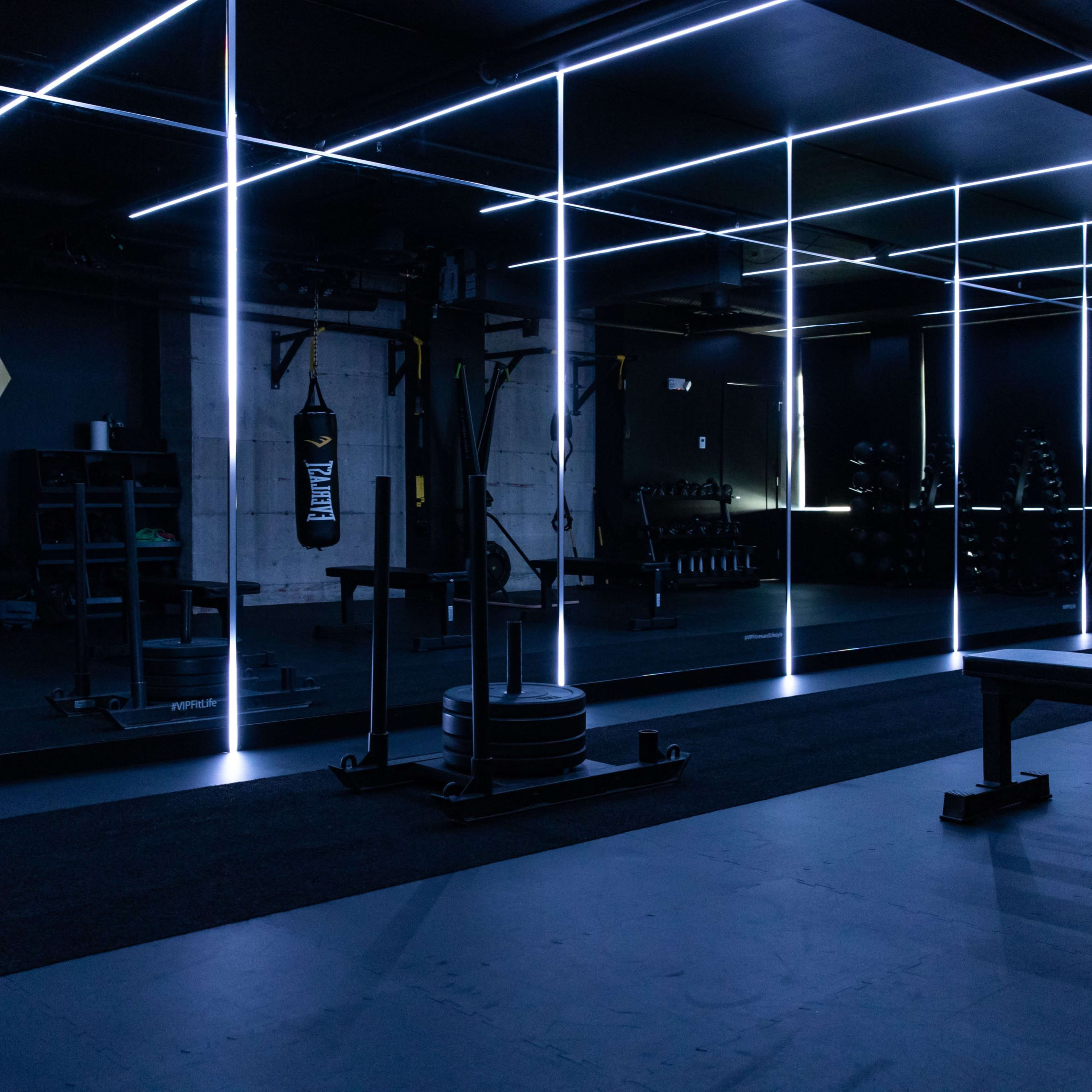 A modern gym features dark walls, illuminated lines, weightlifting equipment, and a mirrored area.