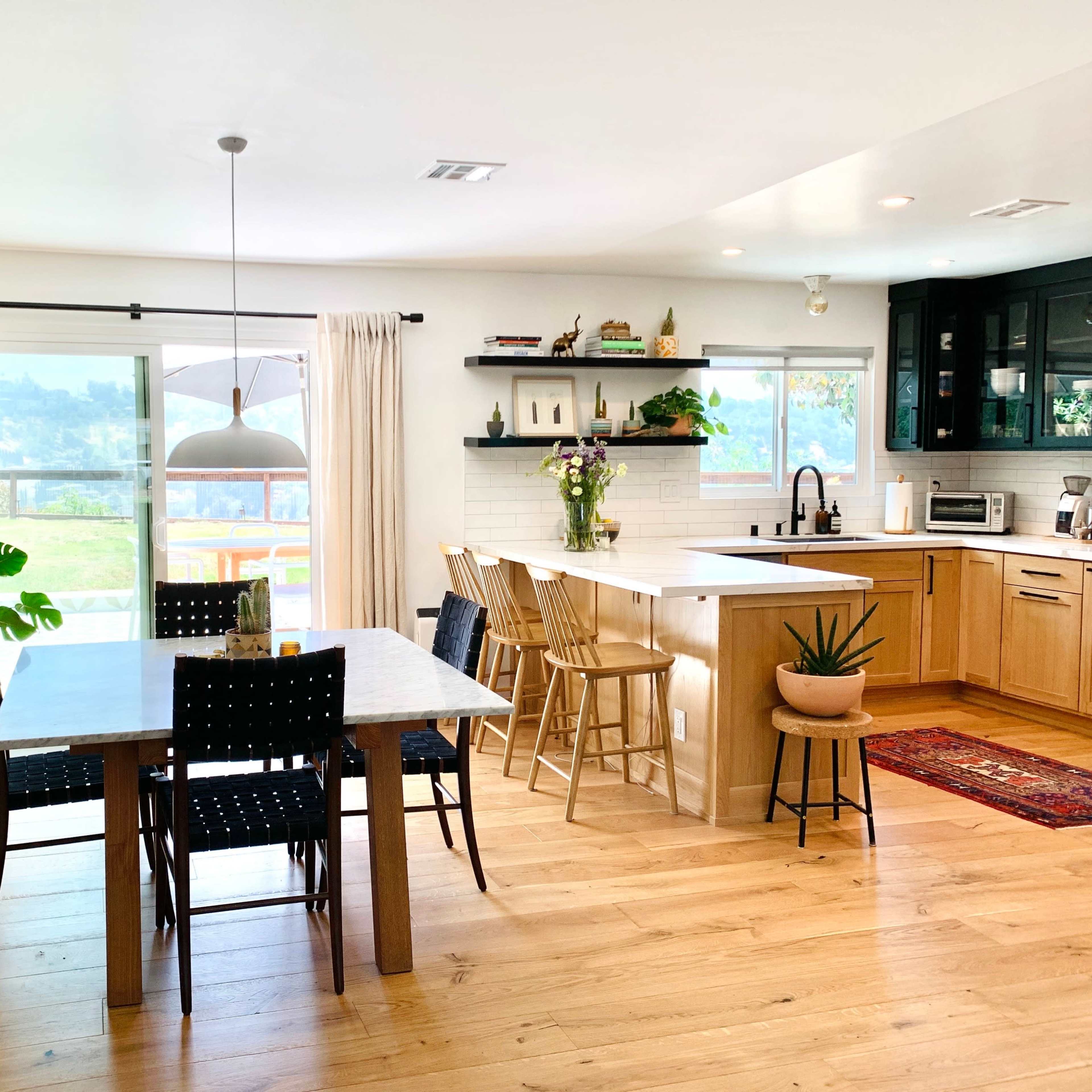 A modern kitchen and dining area with wooden flooring, a dining table with black chairs, and a kitchen featuring light wood cabinets and a large window overlooking a landscape.