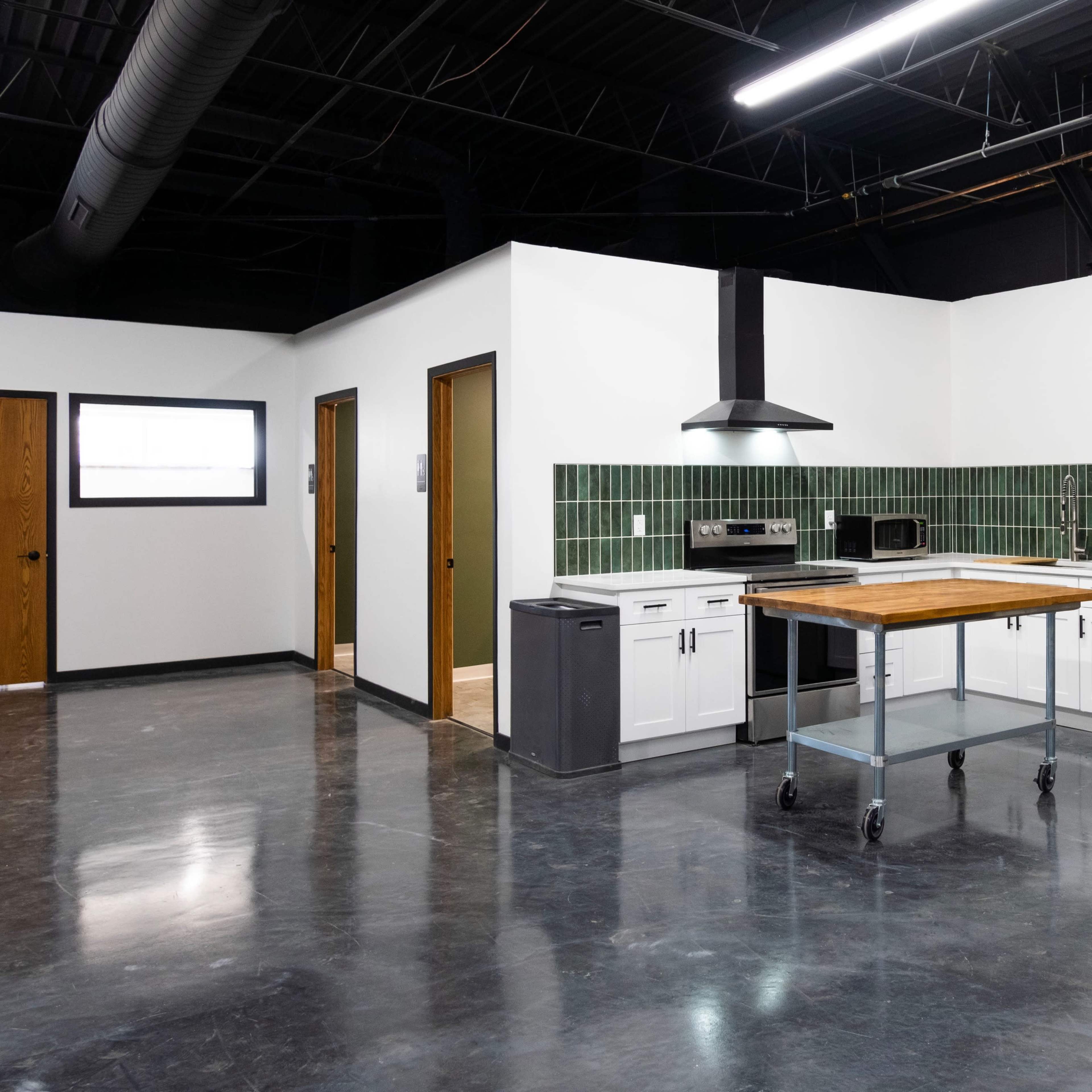 A modern kitchen with stainless steel appliances, a wooden island on wheels, and green tiled backsplash, set in an open space with polished concrete flooring.