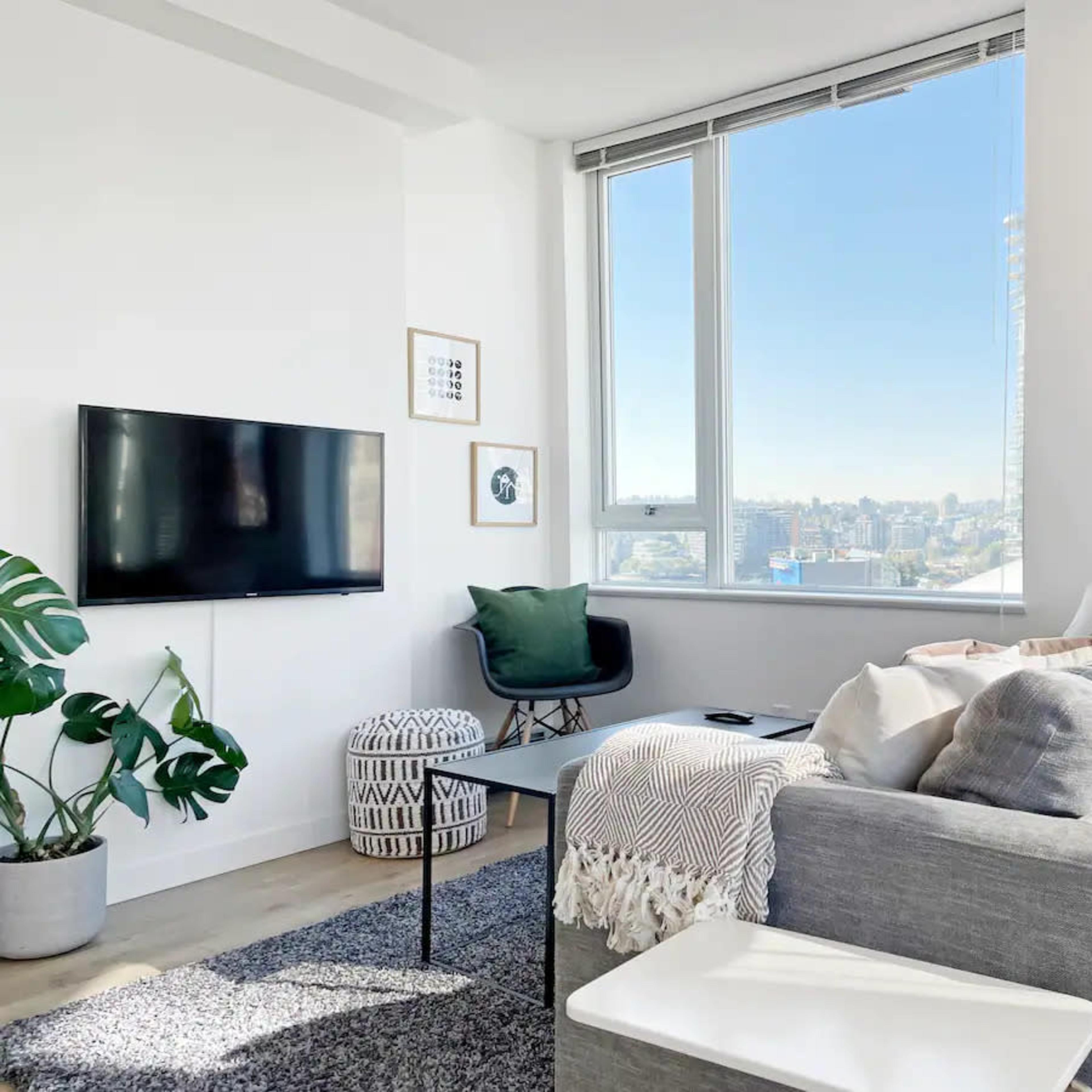 A modern living room features a gray sectional sofa, a black coffee table, a wall-mounted television, and large windows that provide a view of the city skyline.