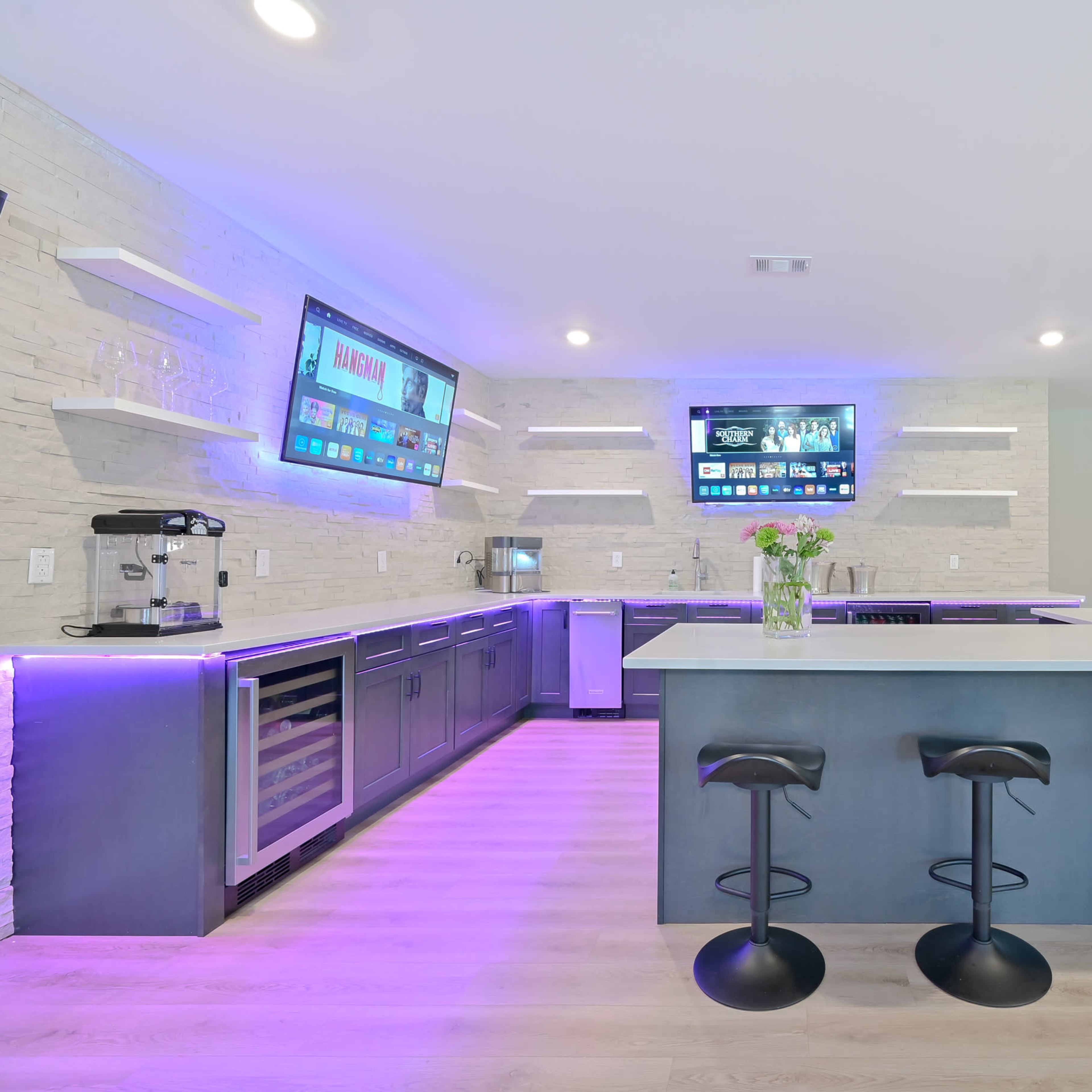 A modern kitchen with a large island, bar seating, and wall-mounted screens displaying information, illuminated with purple lighting.