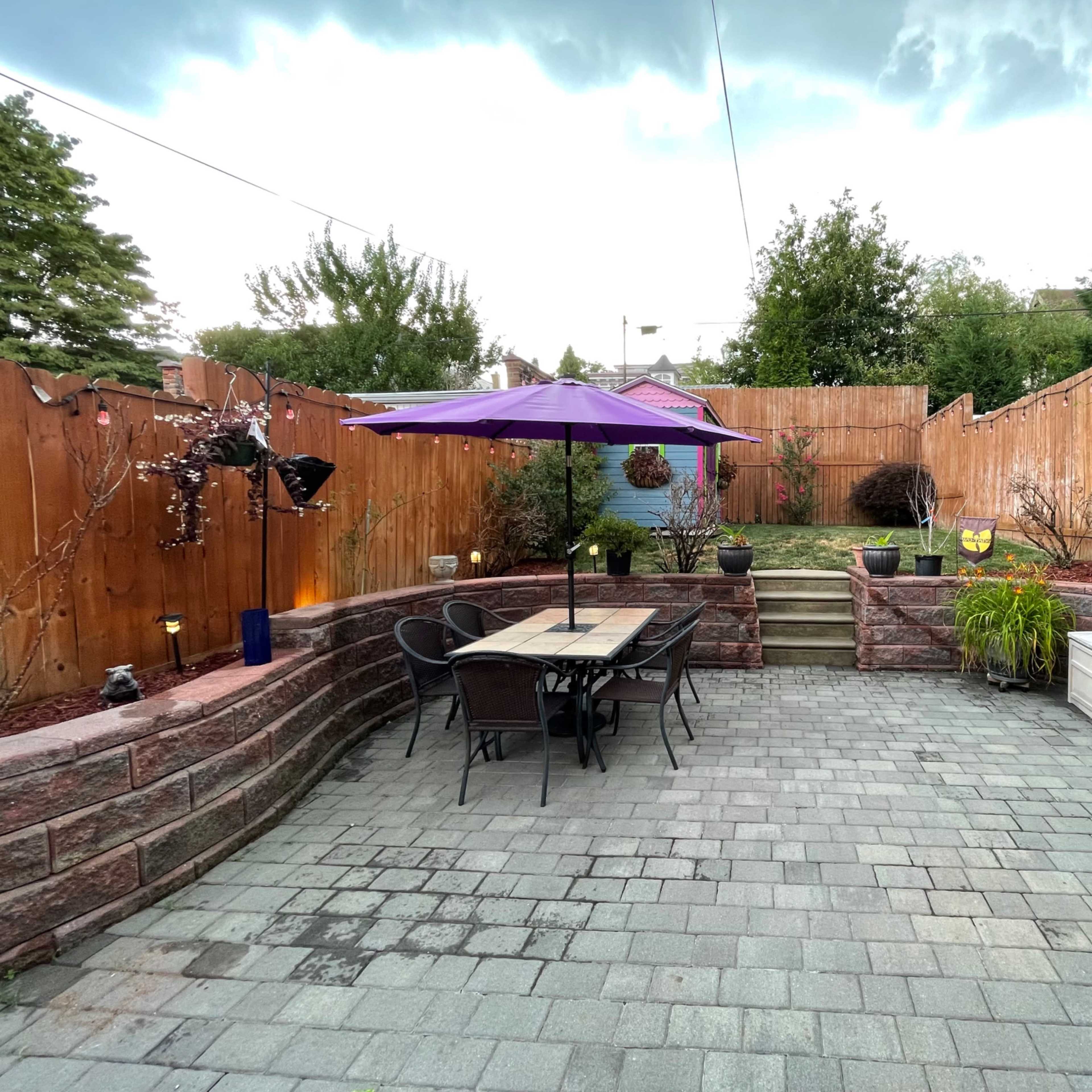 A paved backyard patio features a table and chairs under a purple umbrella, surrounded by garden plants and a wooden fence.
