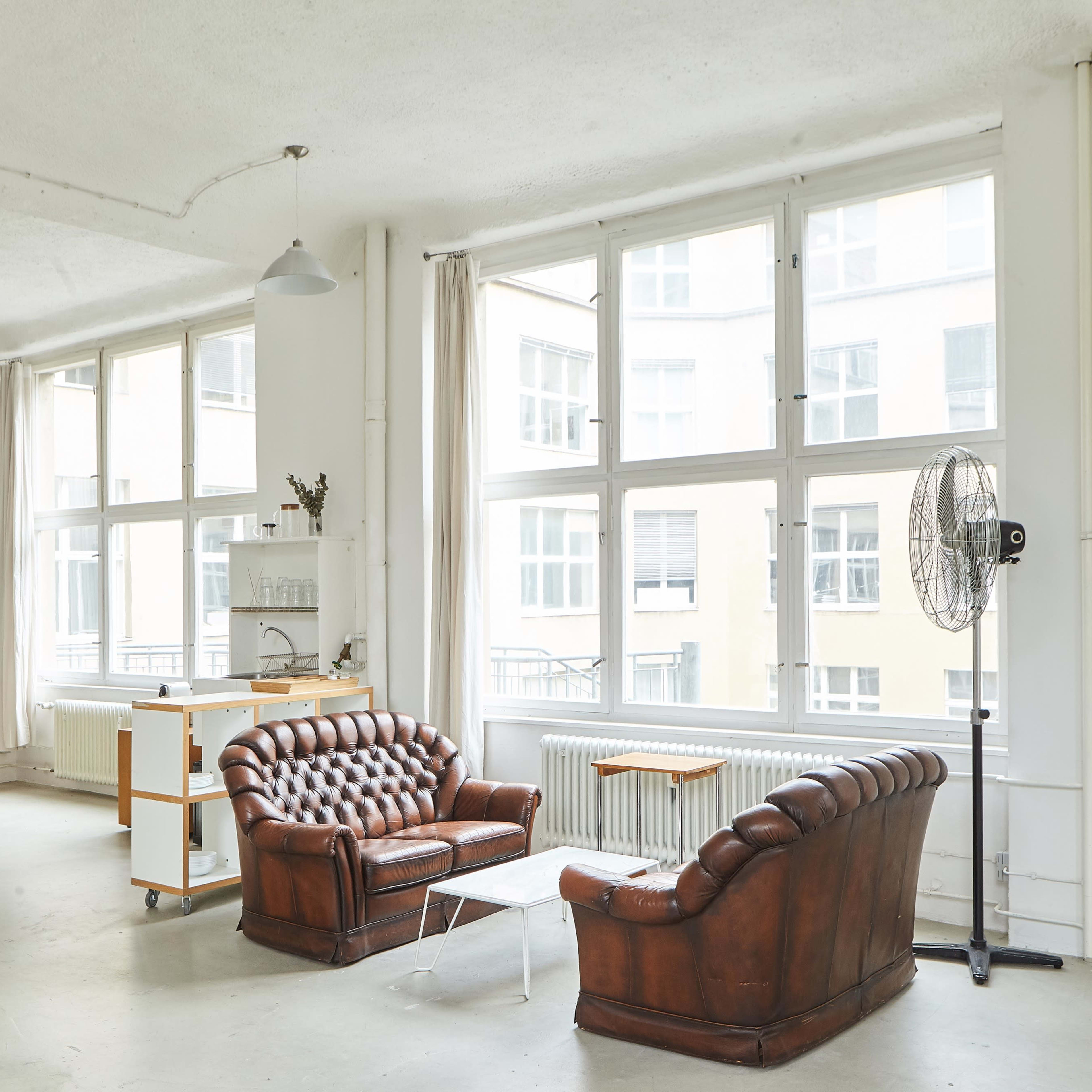 A spacious, brightly lit room features two vintage leather sofas, a small glass table, and large windows overlooking a building.