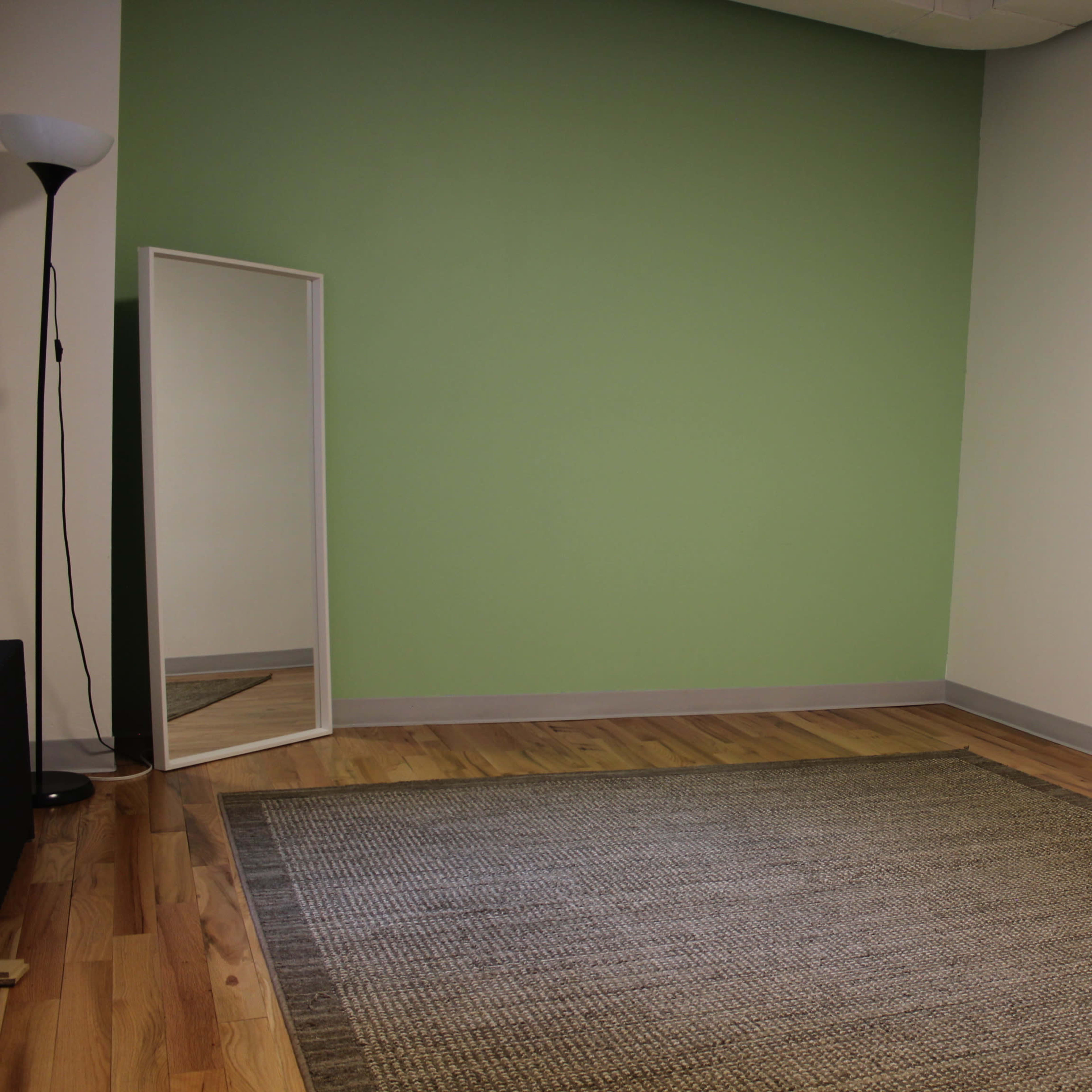 A sparse room with a green wall, wooden flooring, a rectangular rug, a full-length mirror, and a floor lamp.