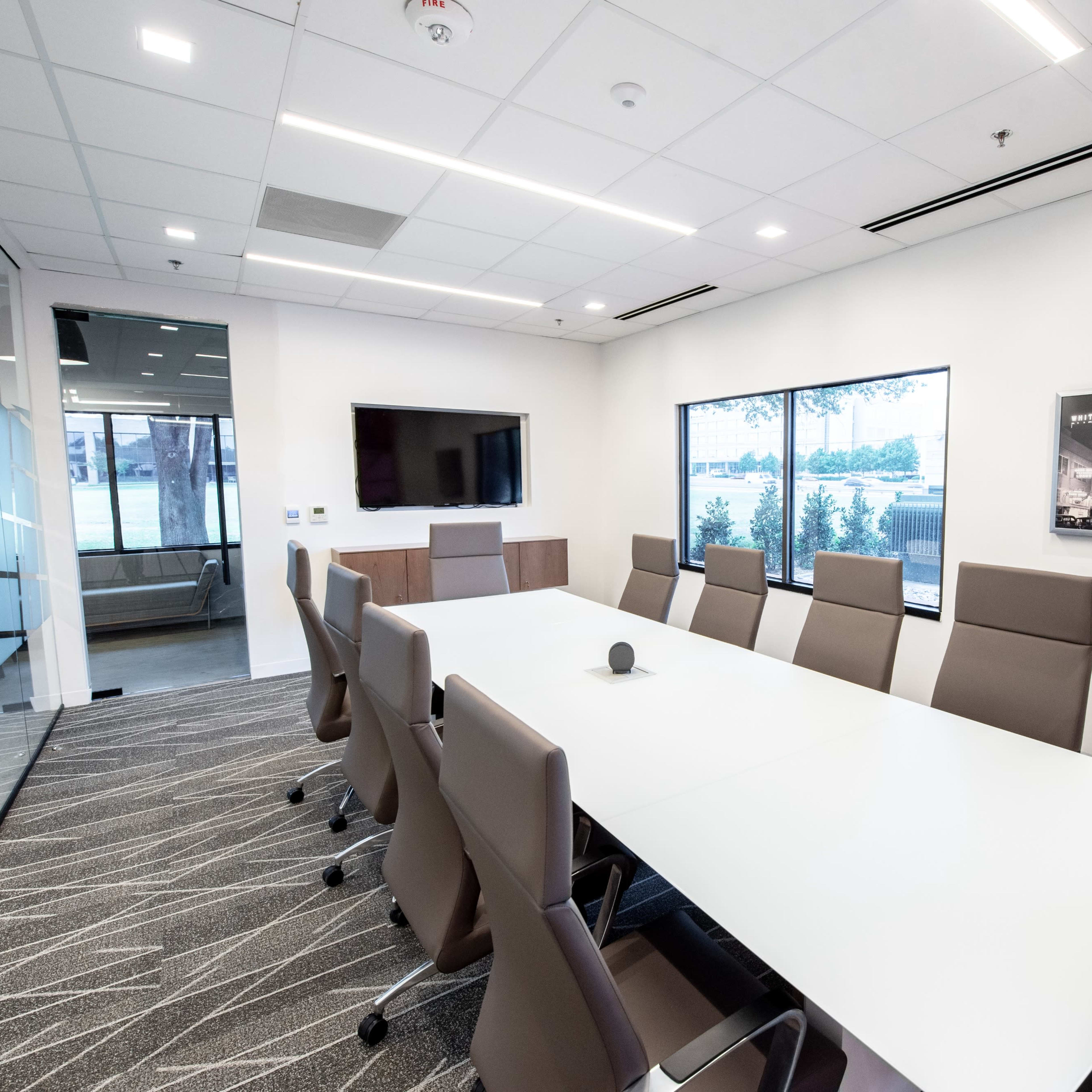 A modern conference room features a large white table surrounded by ergonomic chairs, with a television and large windows showcasing a view outside.