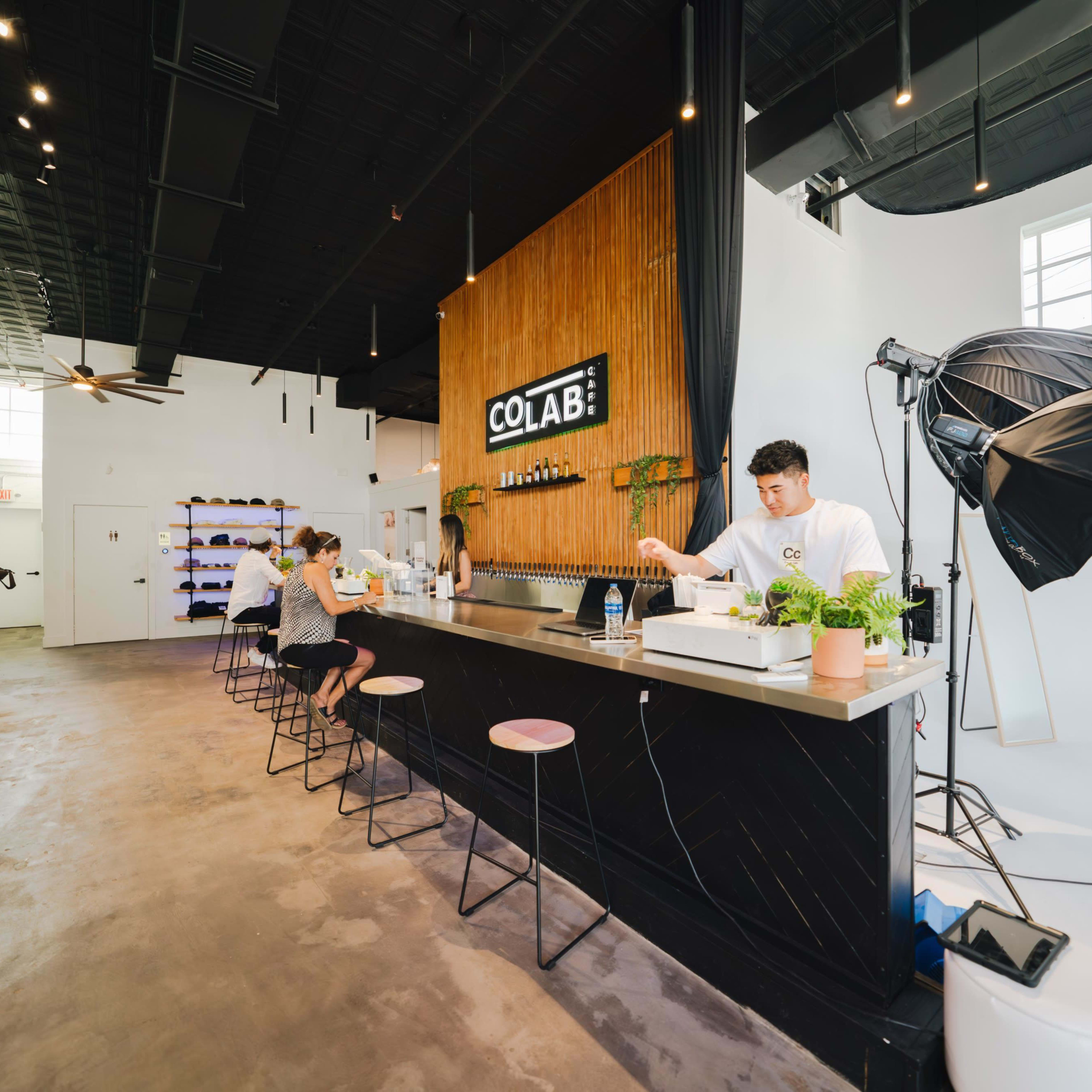 A modern workspace featuring a bar area with stools, a counter with a person attending to customers, and a backdrop labeled "COLAB."
