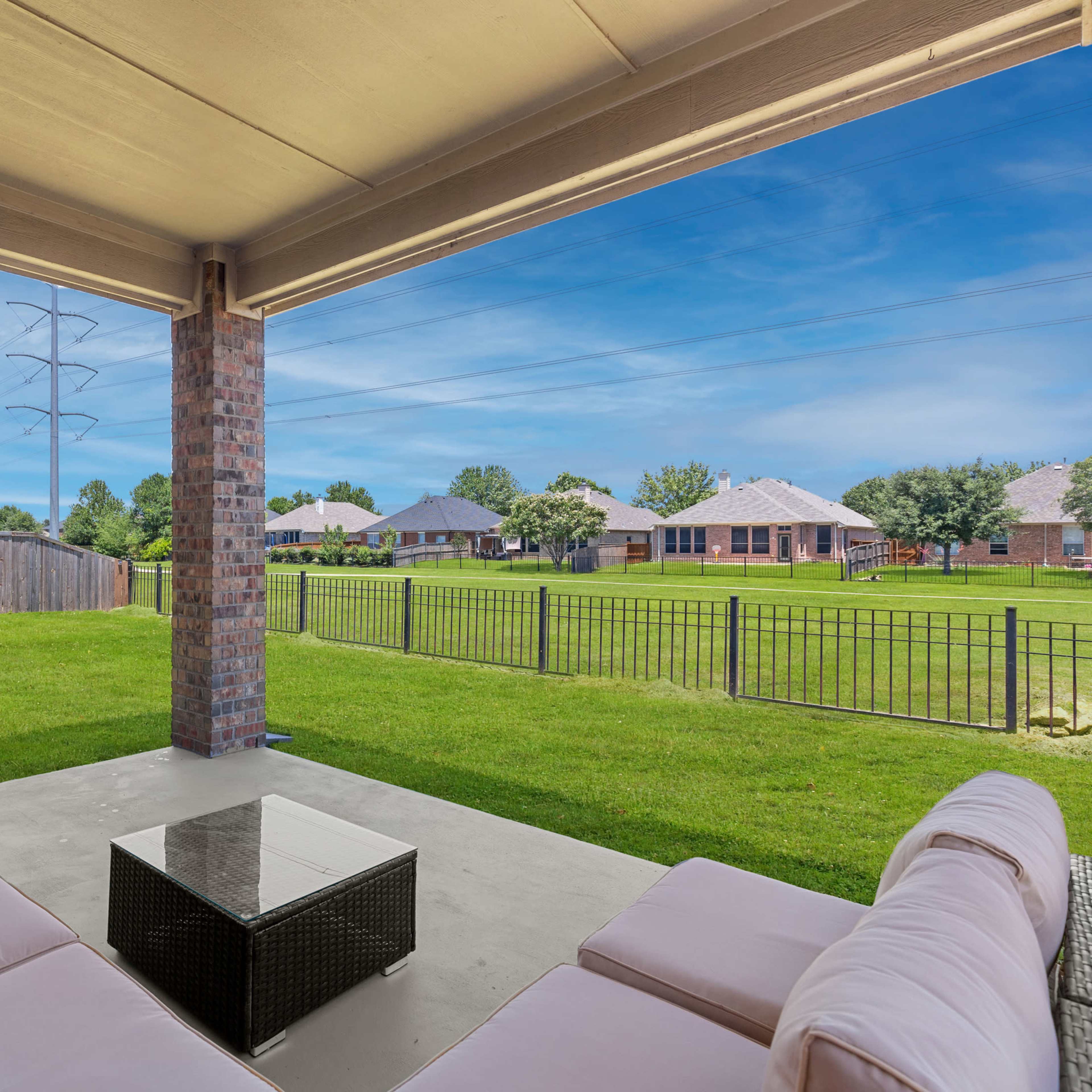 A covered patio with a sectional sofa and coffee table overlooks a green yard and neighboring houses.