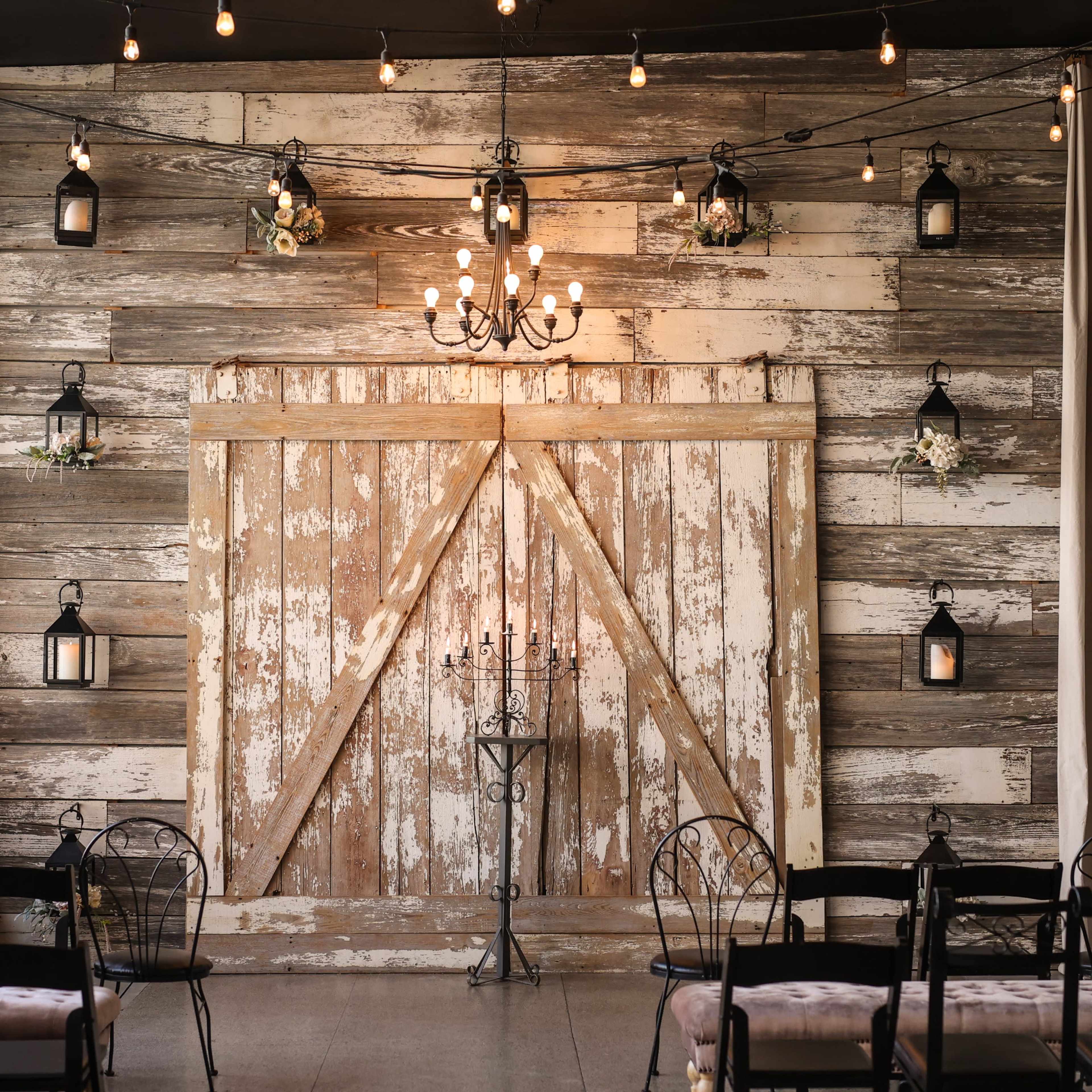 A rustic interior space with a weathered wooden wall and a large barn door, illuminated by hanging lights and featuring decorative lanterns.