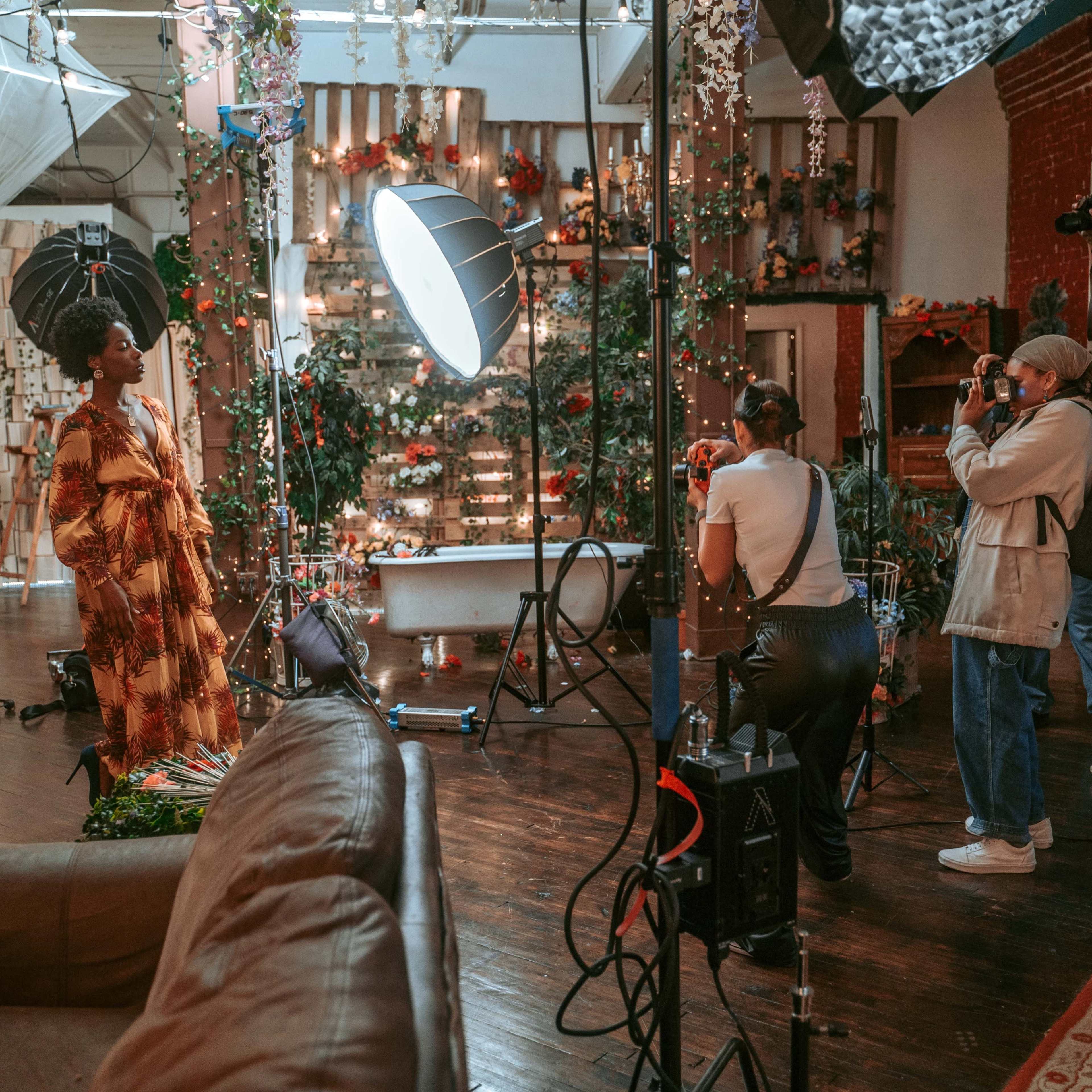 A model poses for a photoshoot in a decorated studio while photographers and crew members adjust equipment and capture images.