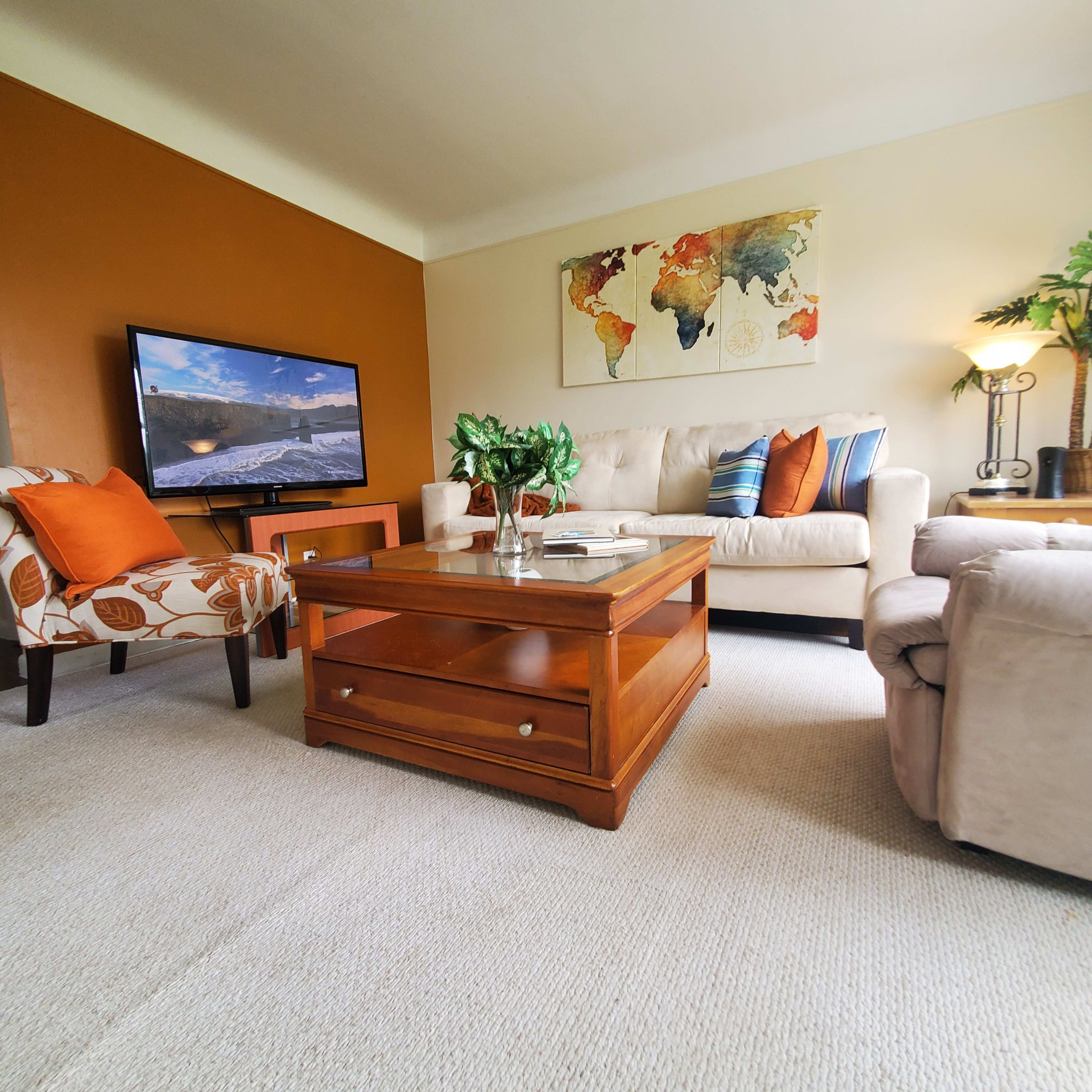 The living room features a cream-colored couch, an armchair with a floral pattern, a wooden coffee table, and a television displaying a scenic image against a backdrop of warm-colored walls.