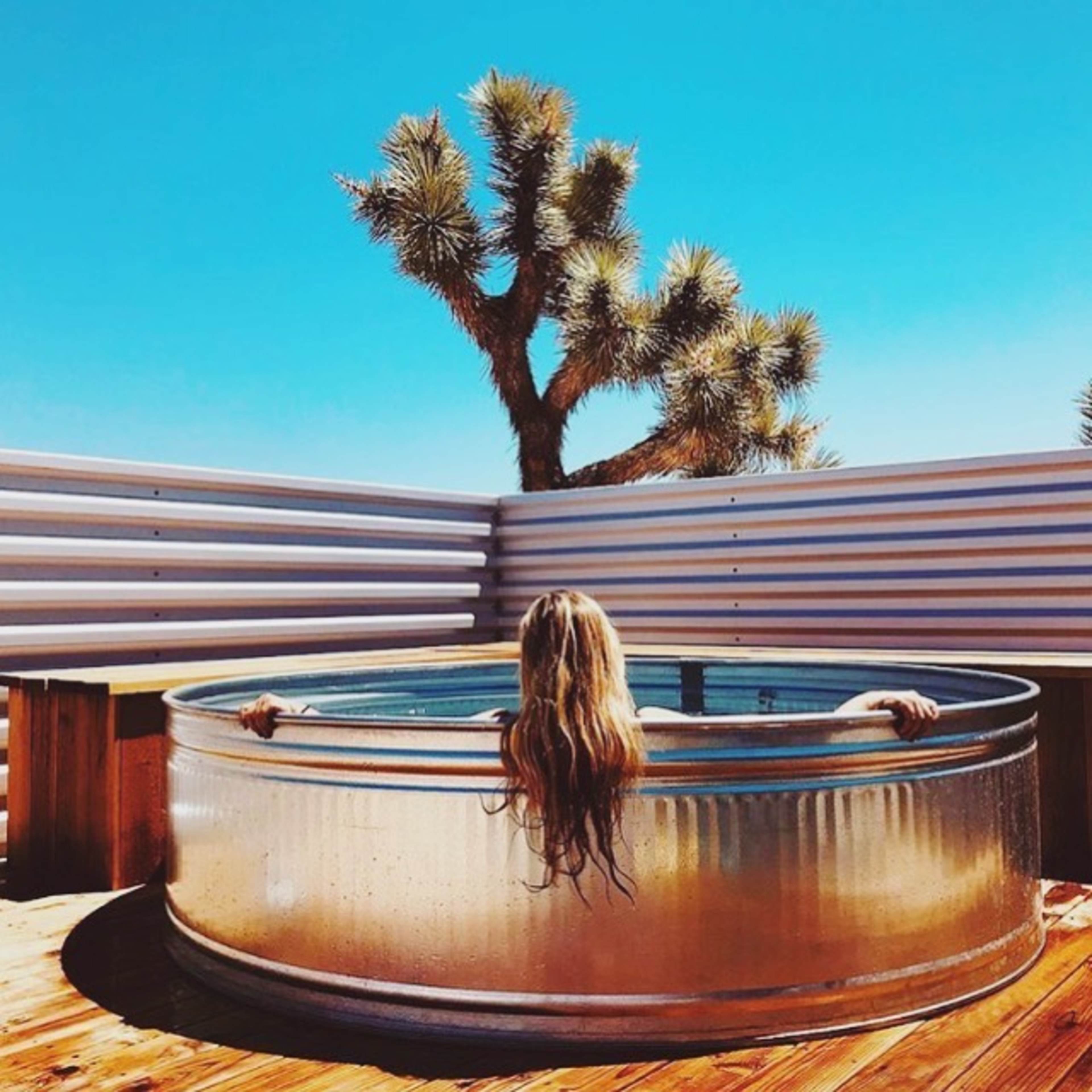 A person with long hair relaxes in a circular metallic tub on a wooden deck, with a Joshua tree visible in the background under a clear blue sky.