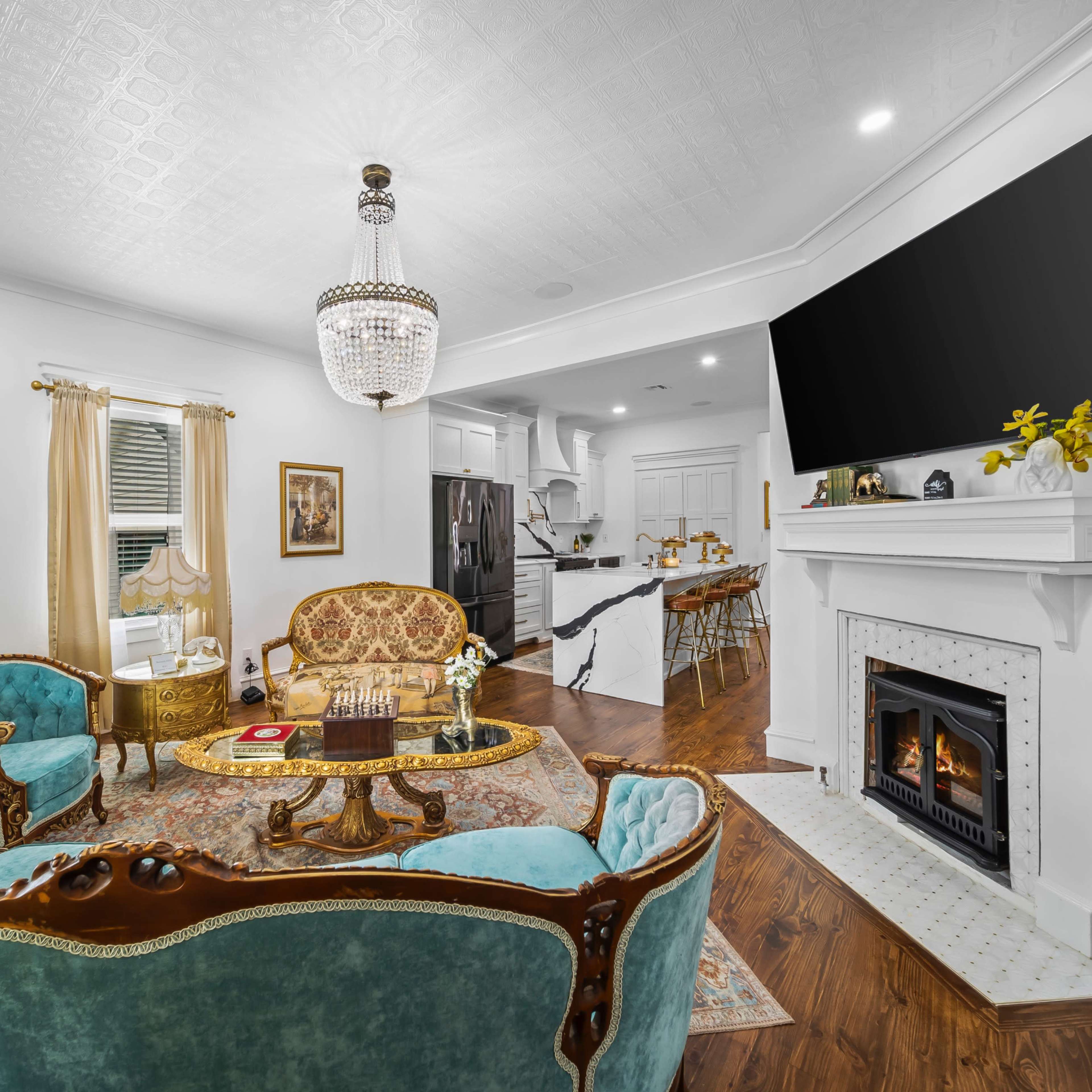 A bright living room features turquoise upholstered chairs, a coffee table, a fireplace, and a mounted television, with a kitchen visible in the background.