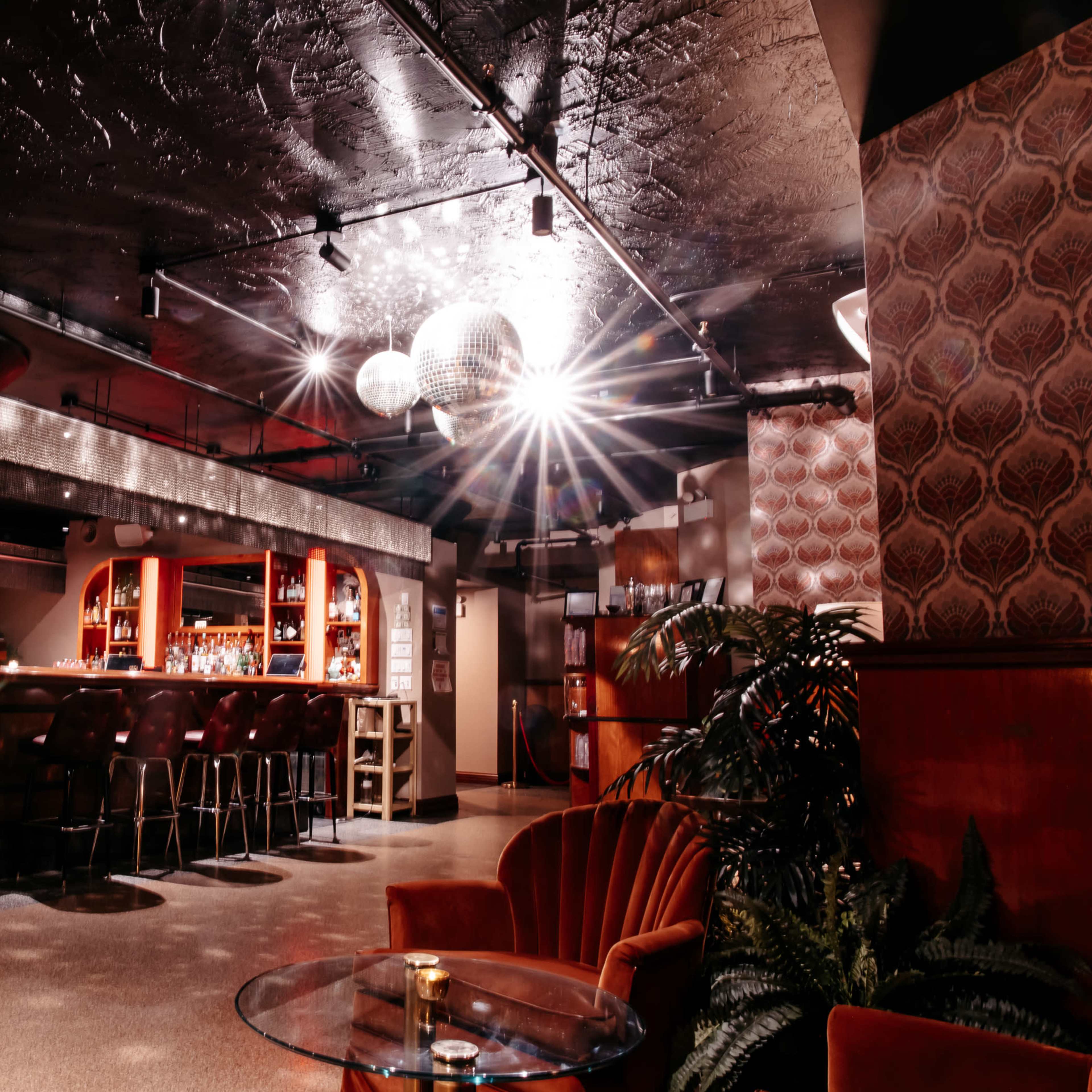 A dimly lit bar features a circular counter with high stools, surrounded by retro decor and a large disco ball reflecting light.