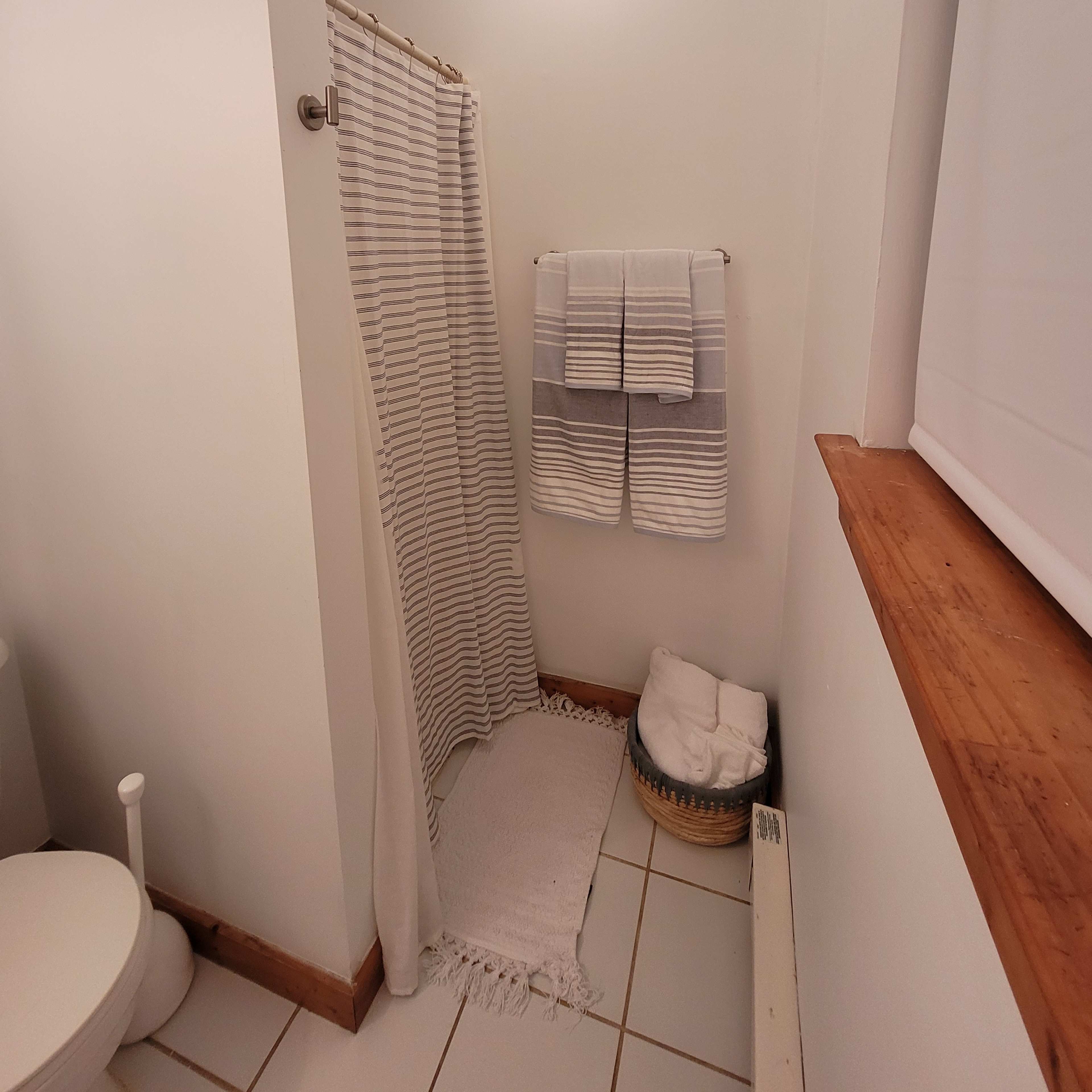 A bathroom with a shower area, a toilet, a woven basket containing towels, and neatly arranged bath linens hanging on the wall.