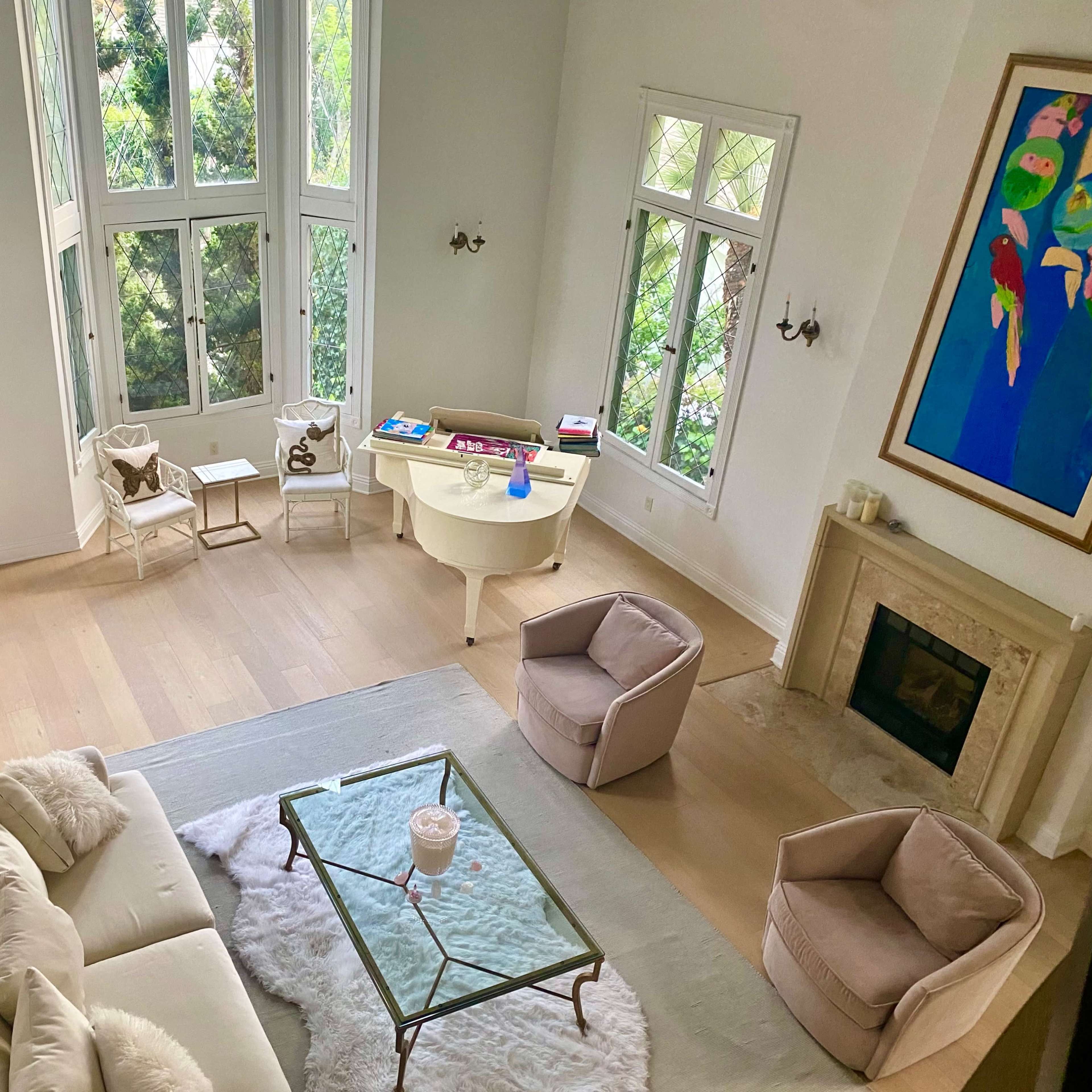 A spacious living room features large windows, a glass coffee table on a light rug, and soft seating arranged around the fireplace.