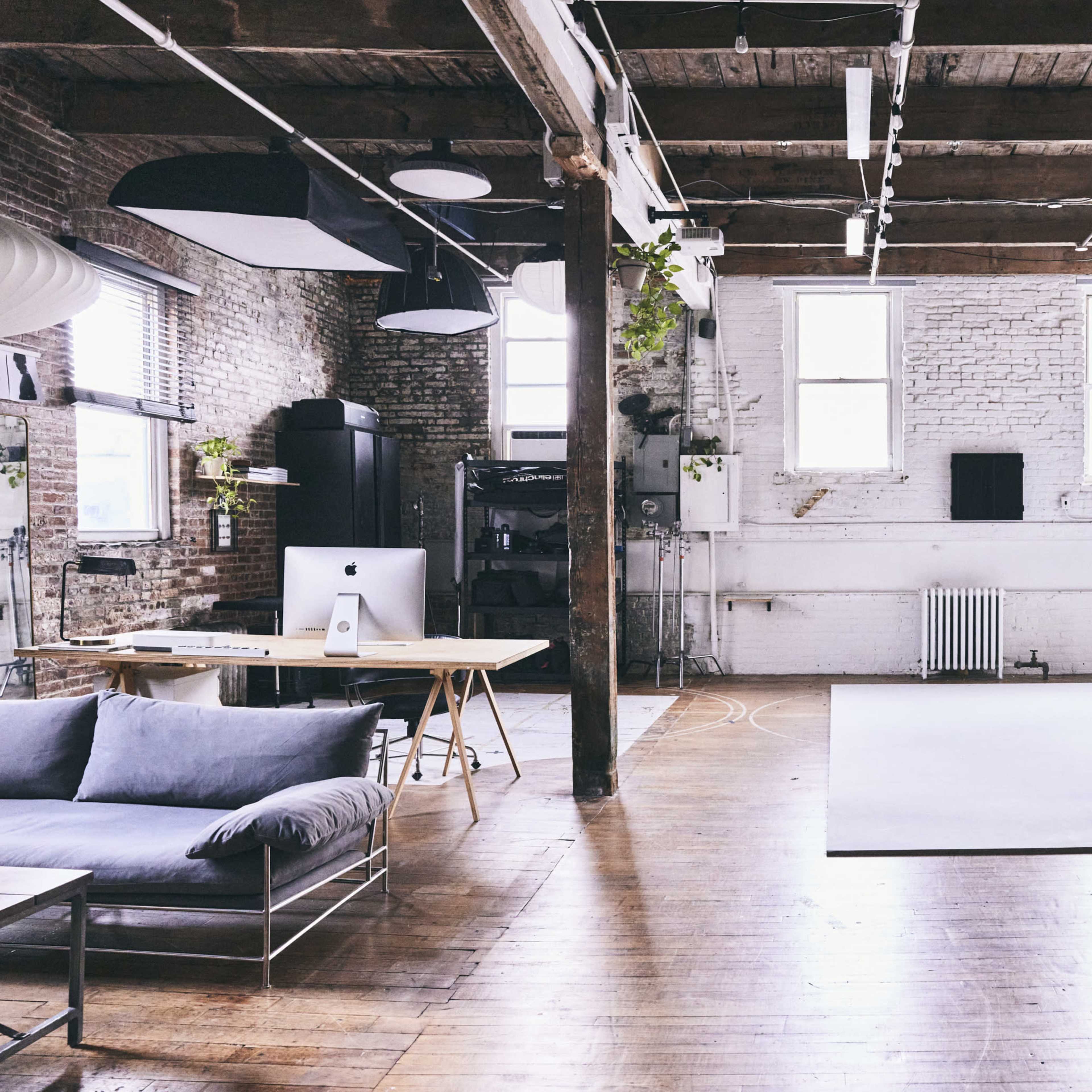 A spacious, well-lit studio with wooden floors, exposed brick walls, and a large white backdrop area for photography.