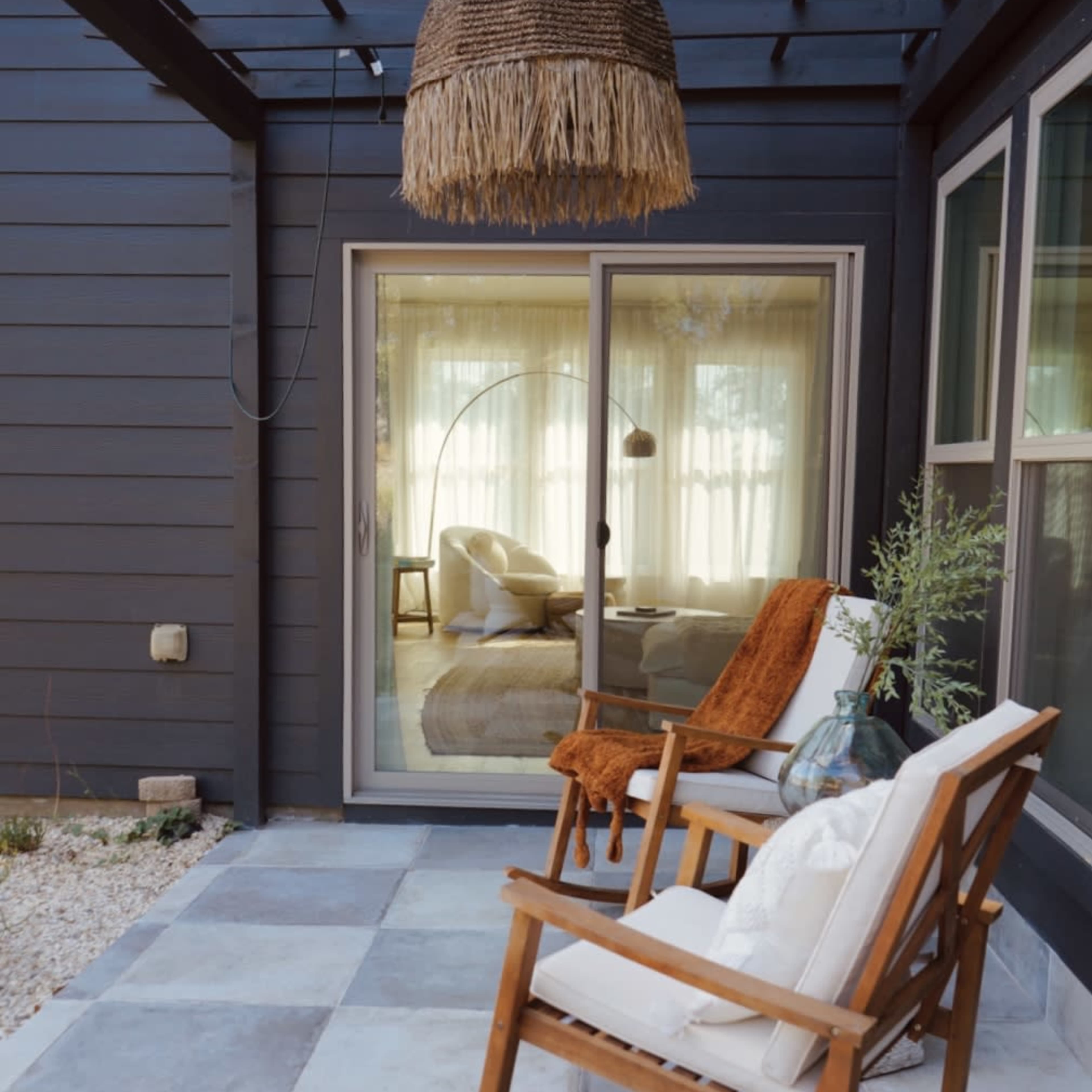 A pair of wooden rocking chairs sit on a stone patio in front of a large glass door leading to a light-filled indoor space.