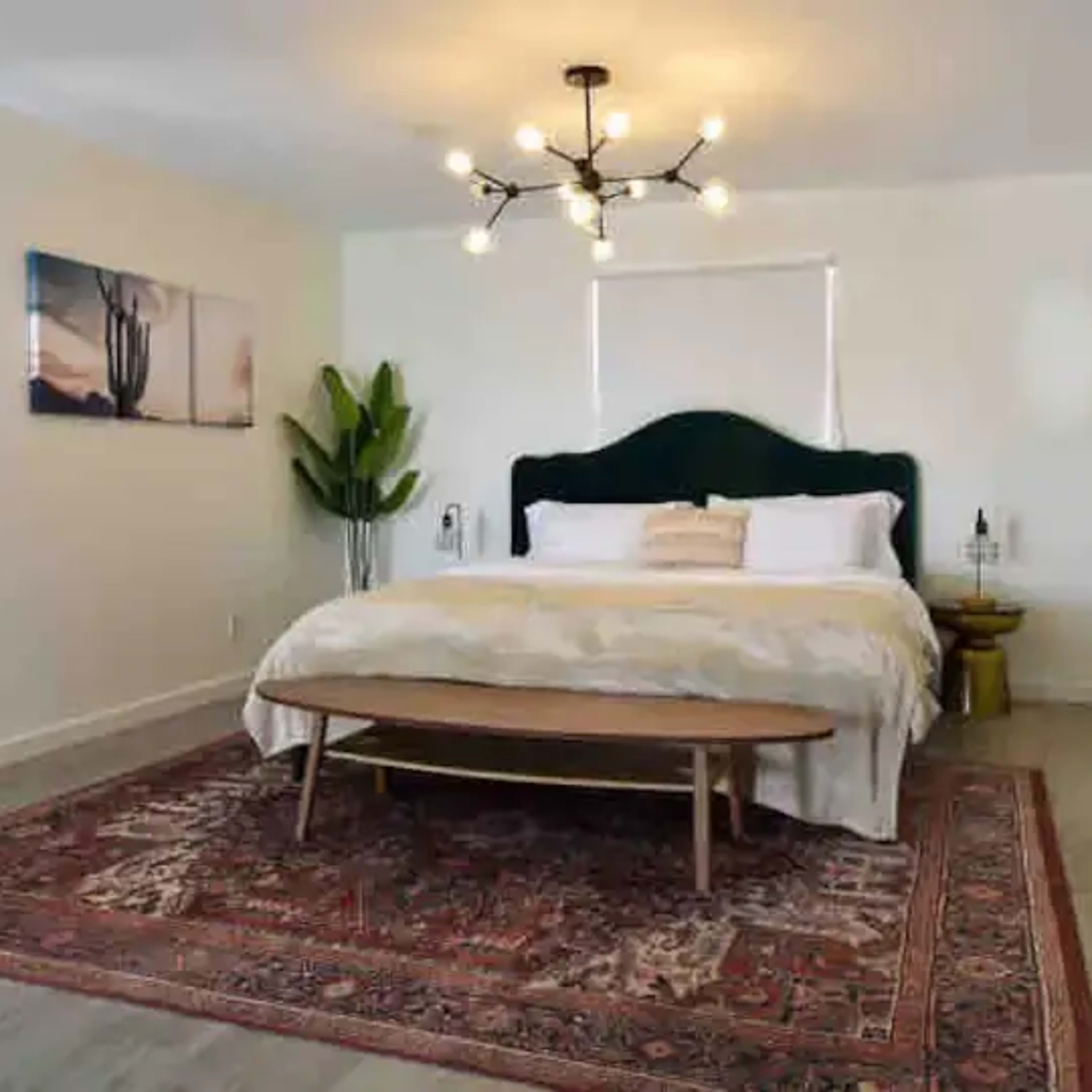 A modern, well-lit bedroom with a large bed, a wooden table in front, and decorative plants.