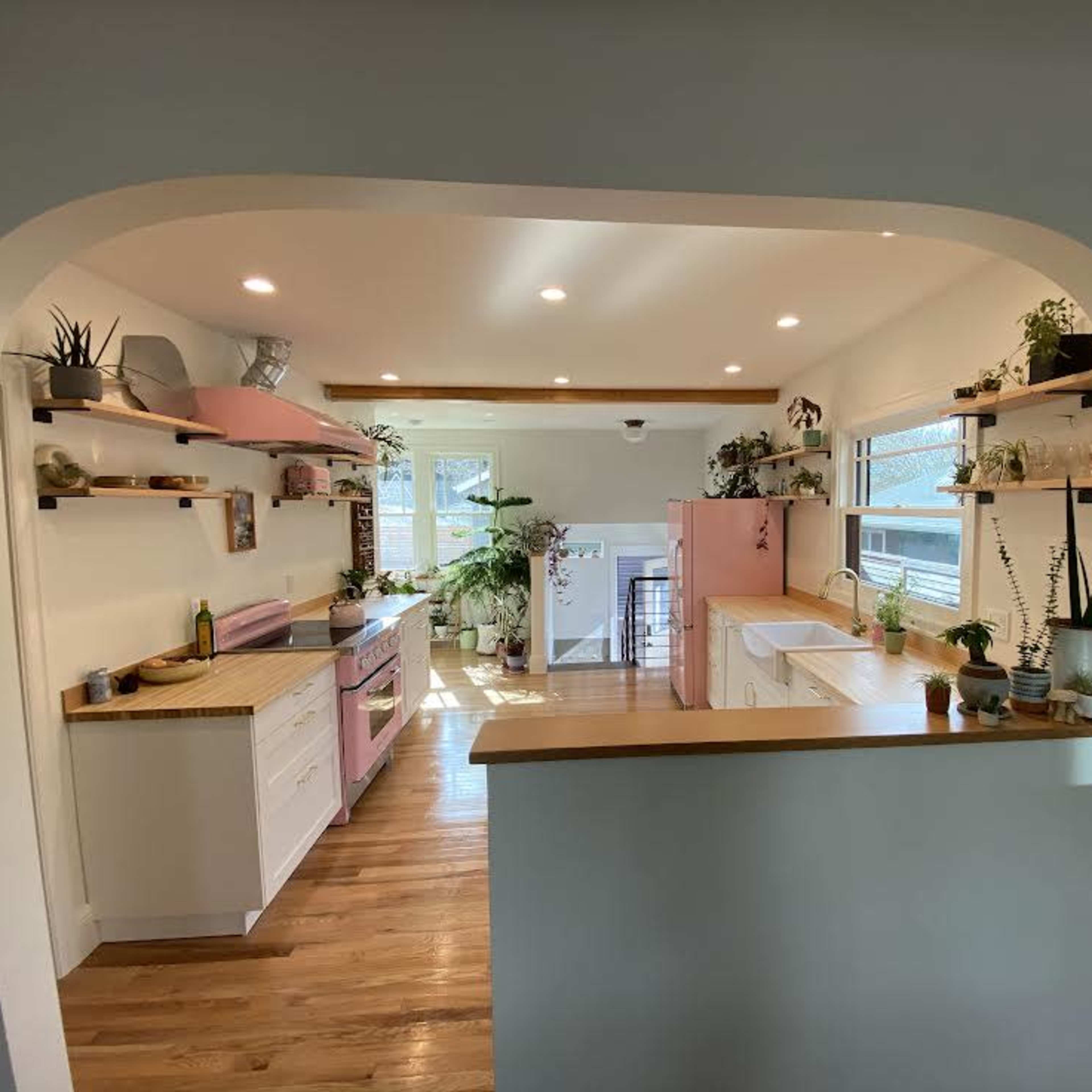 A view of a kitchen featuring pink and white cabinetry, wooden countertops, and ample greenery displayed on shelves.