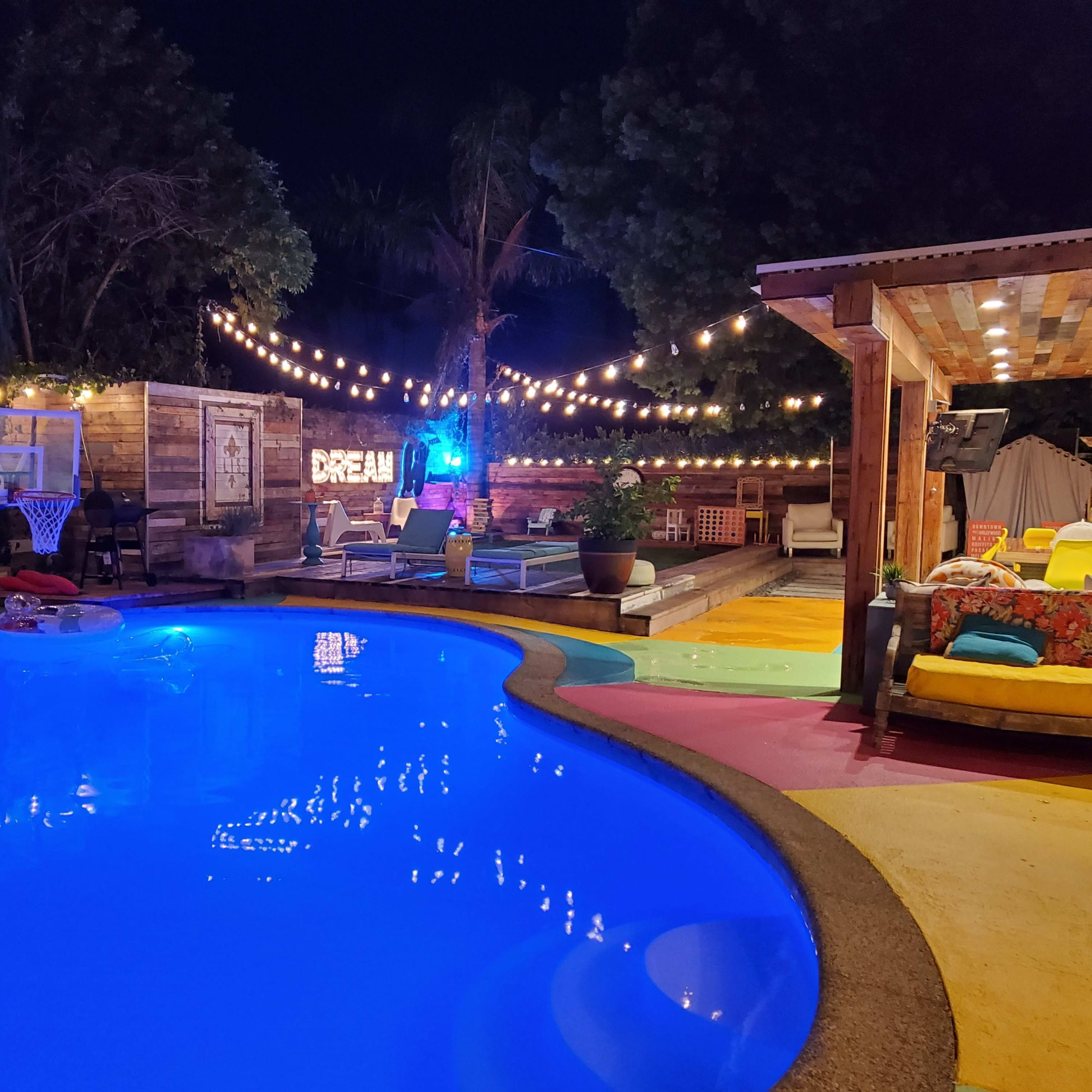 A brightly lit backyard pool area features a curvy pool, a hammock, lounge chairs, and string lights illuminating the space at night.