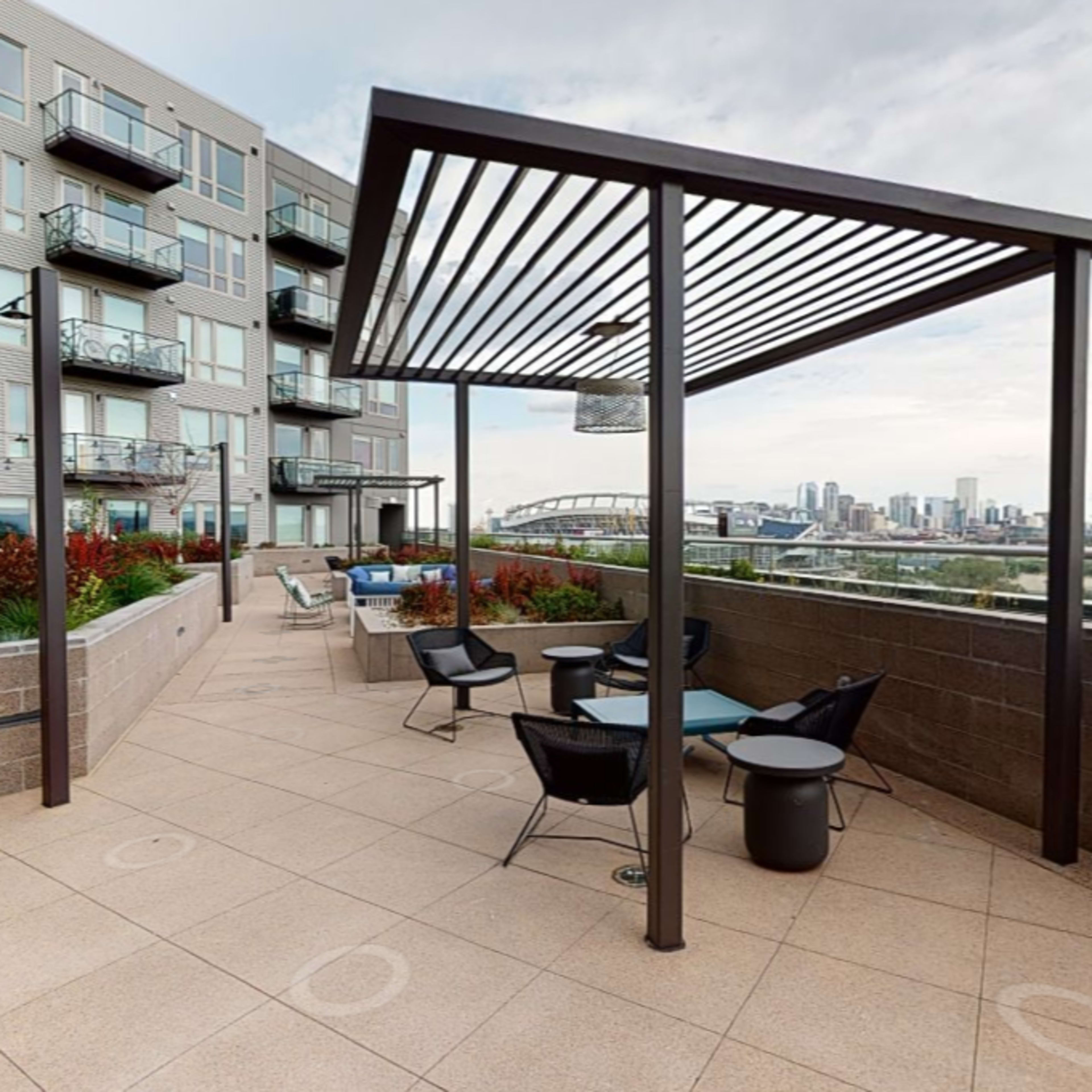 A rooftop terrace with several chairs and tables, surrounded by planters and a view of a city skyline.