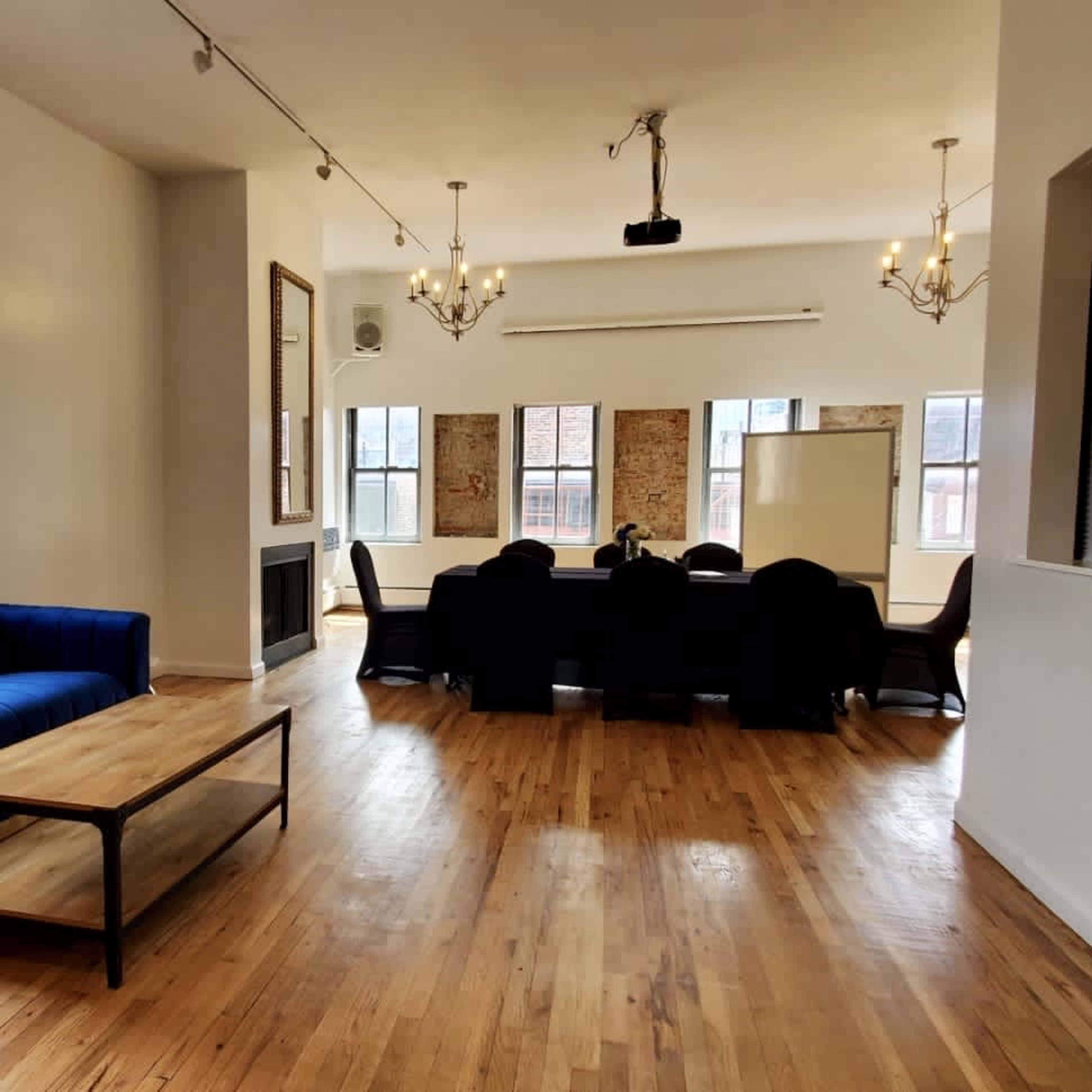 A spacious, well-lit room featuring a blue sofa, a wooden coffee table, and a conference area with a black dining set under chandeliers.