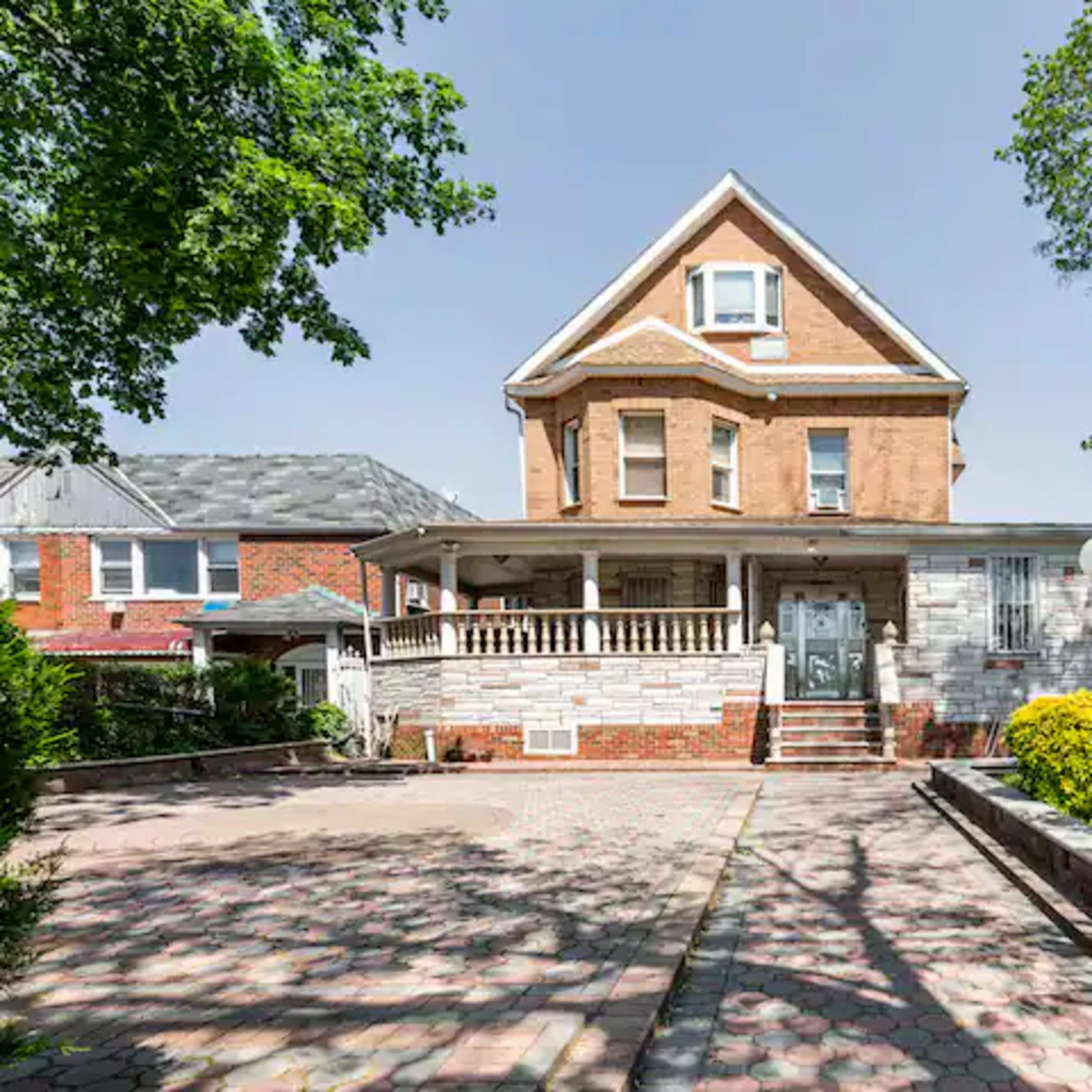 A large two-story brick house with a front porch, surrounded by greenery and a paved driveway.