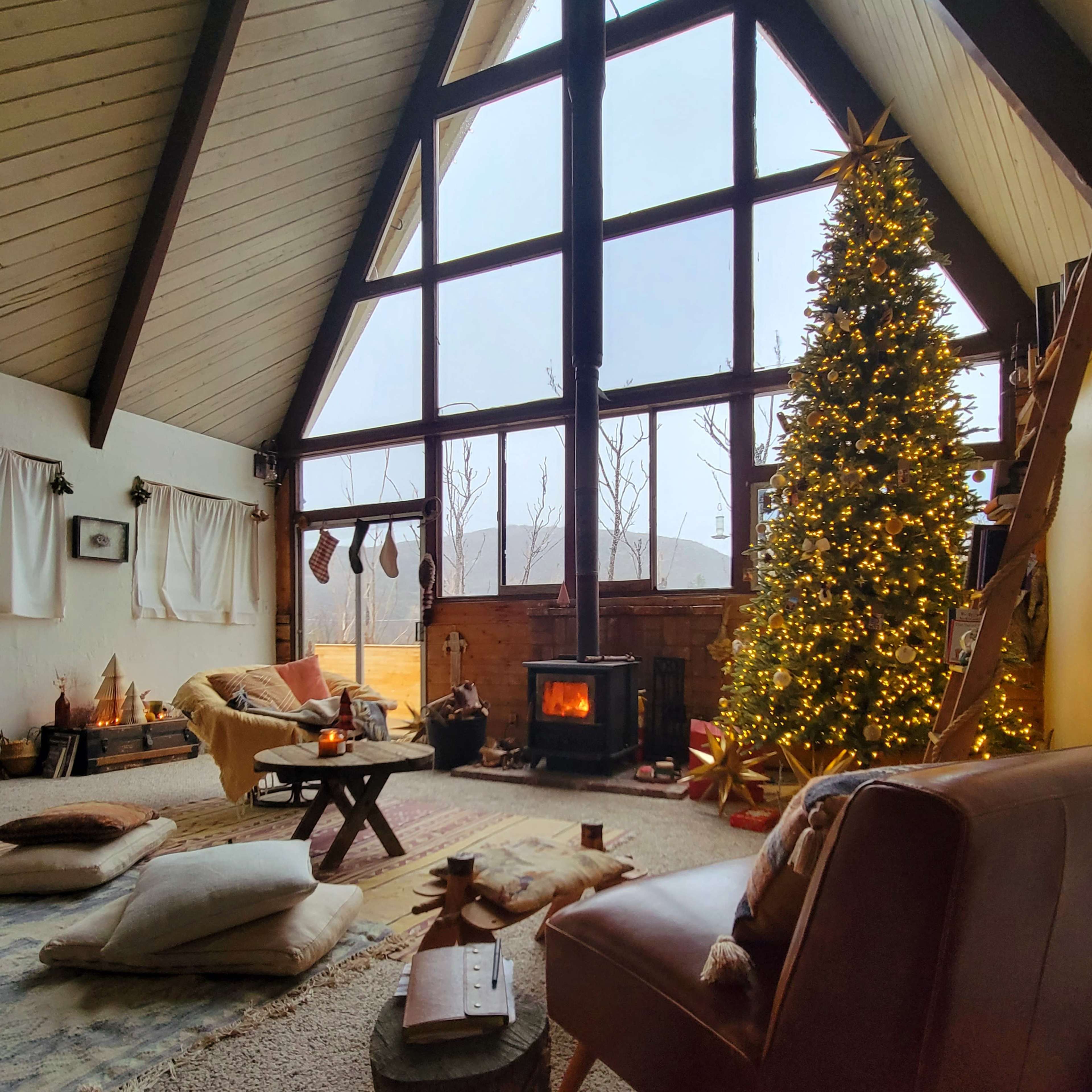 A cozy living room features large windows, a decorated Christmas tree, a fireplace, and comfortable seating.