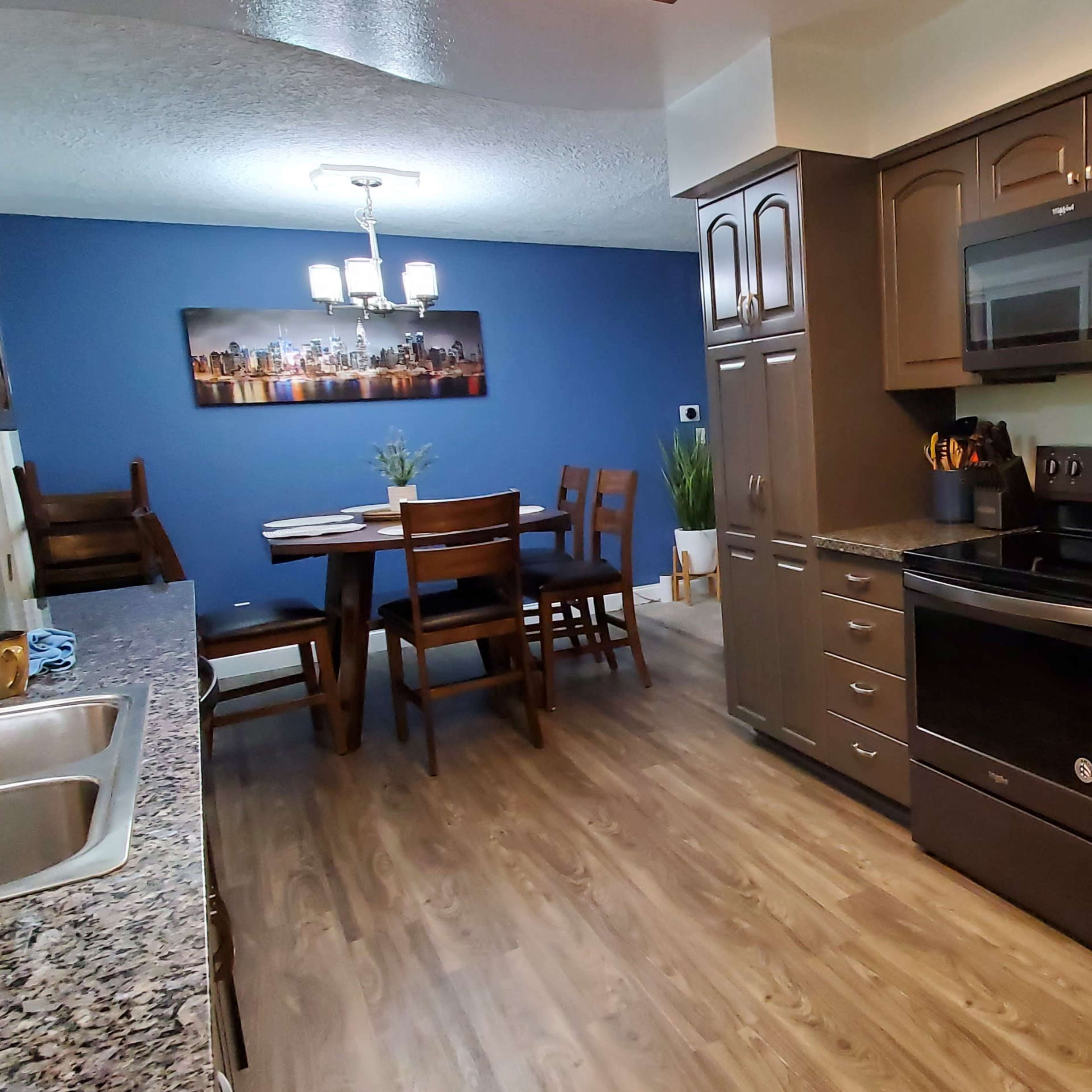 A modern kitchen with dark cabinets, a granite countertop, stainless steel appliances, and an adjacent dining area featuring a wooden table and chairs against a blue wall.