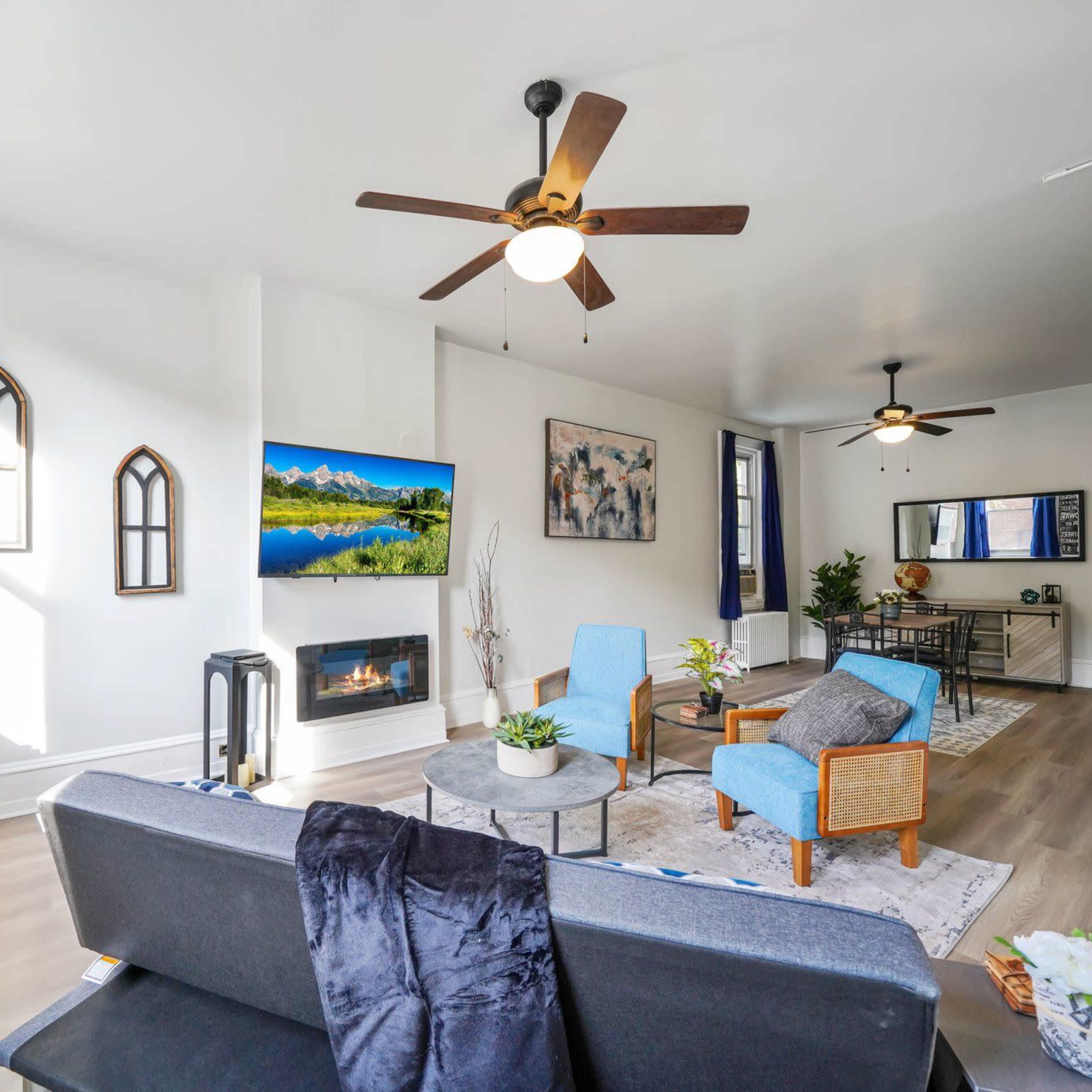 A modern living room features a gray sofa, two blue chairs, a television mounted on the wall, and a decorative fireplace.