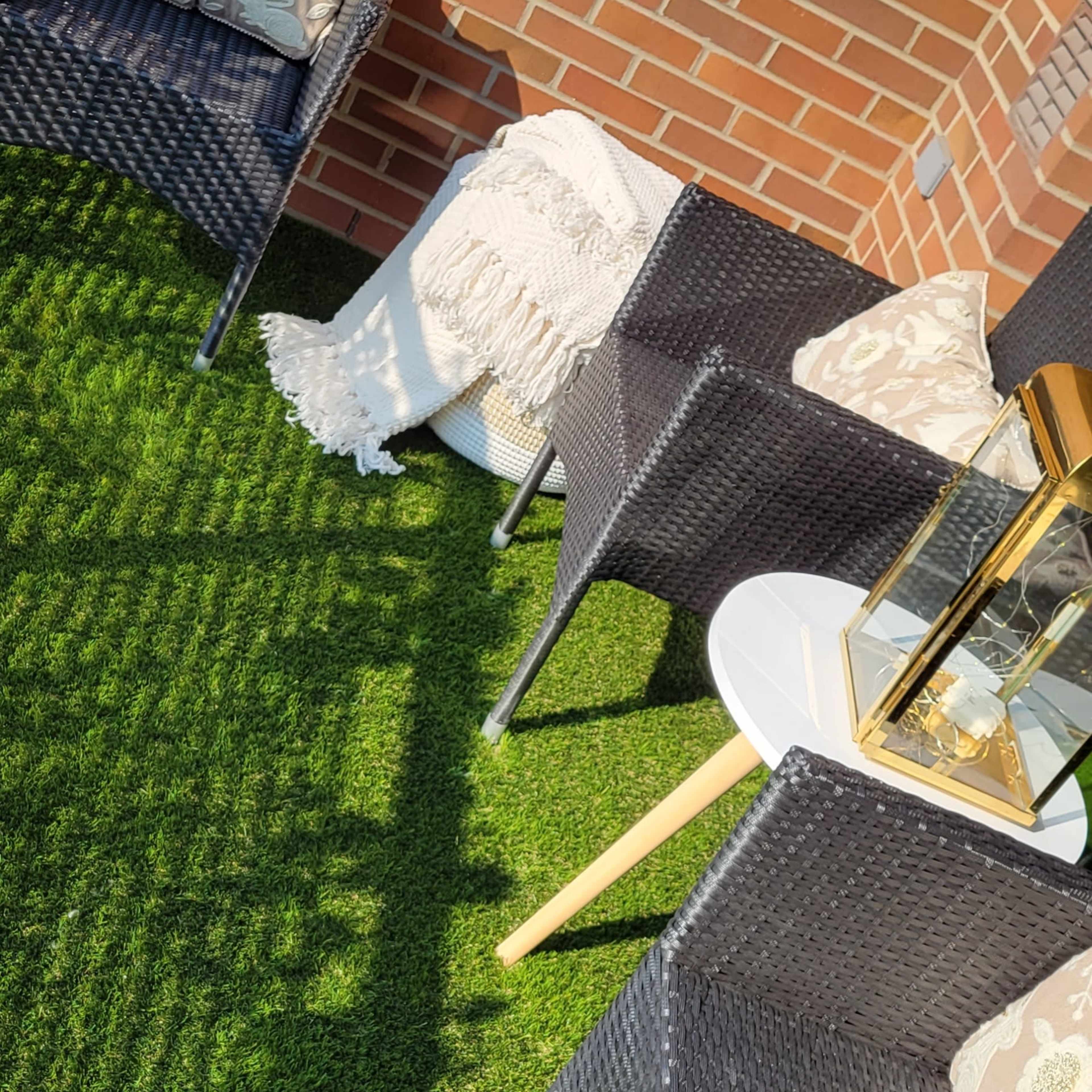 A small outdoor seating area featuring two black wicker chairs with decorative pillows, a side table, and a cozy throw draped over the side, all situated on artificial grass next to a brick wall.