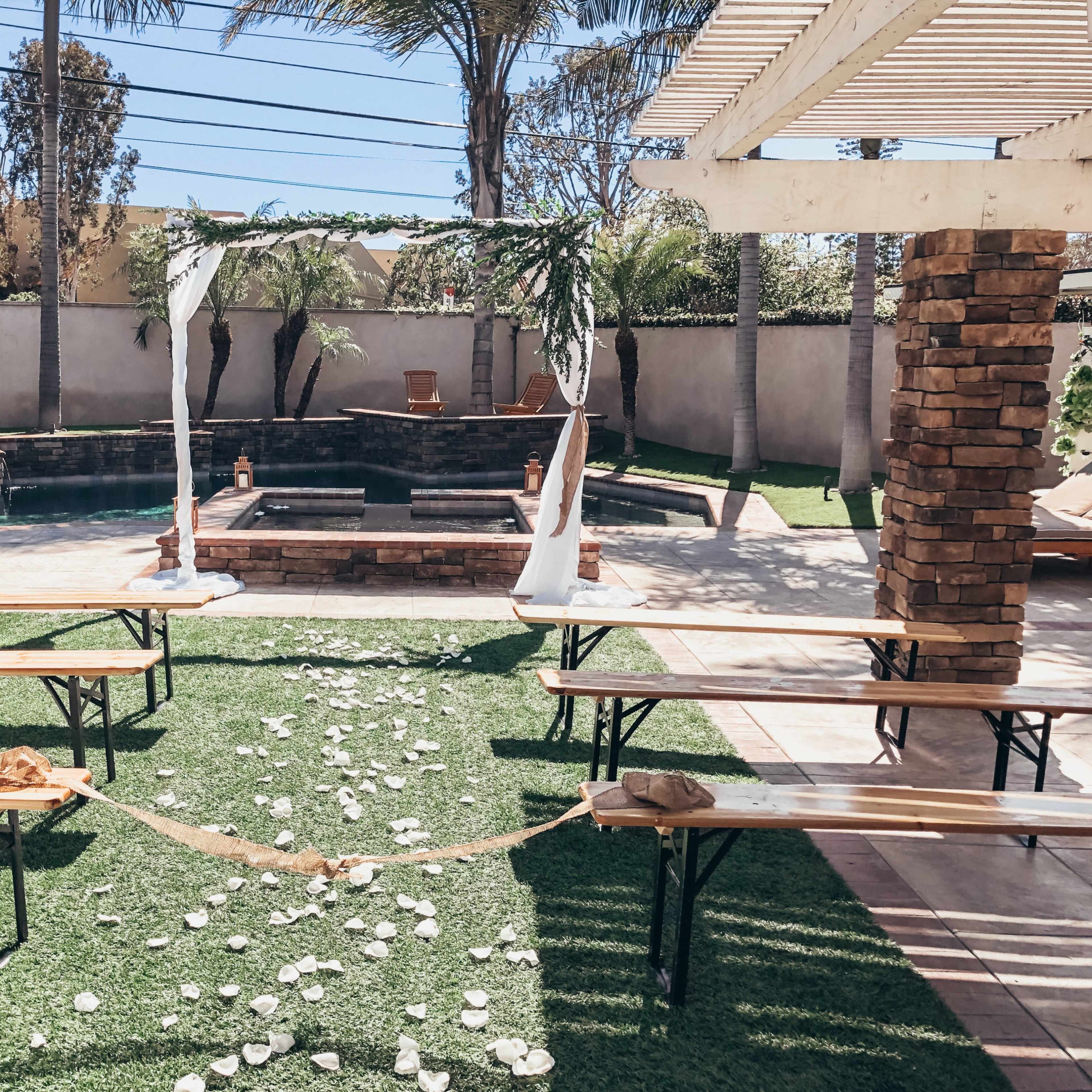 A wedding setup features a stone altar surrounded by benches and a path of scattered flower petals leading to it, all set in a backyard with a swimming pool and palm trees.