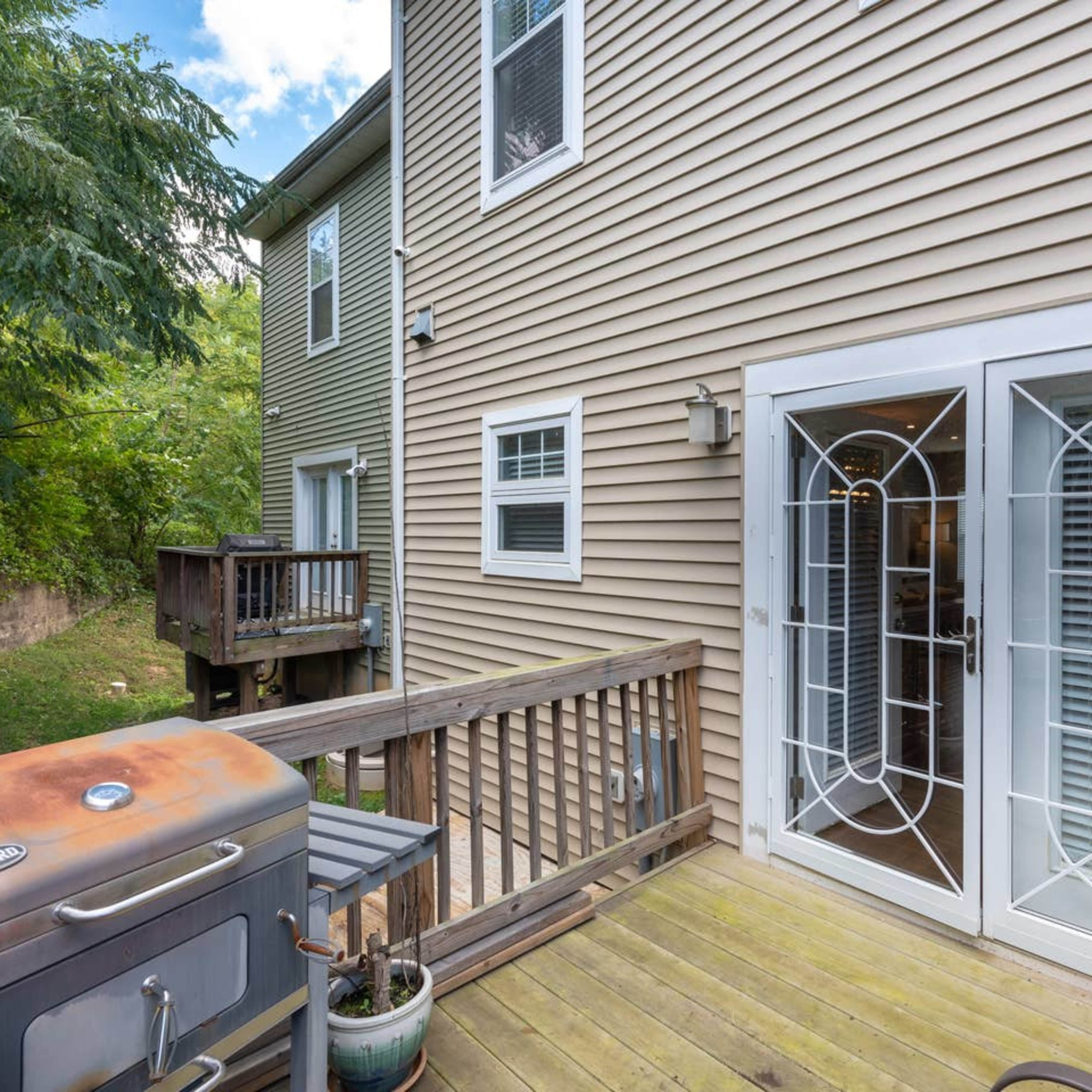 A wooden deck with a metal grill in the foreground, leading to a set of double glass doors on the side of a beige-colored house.