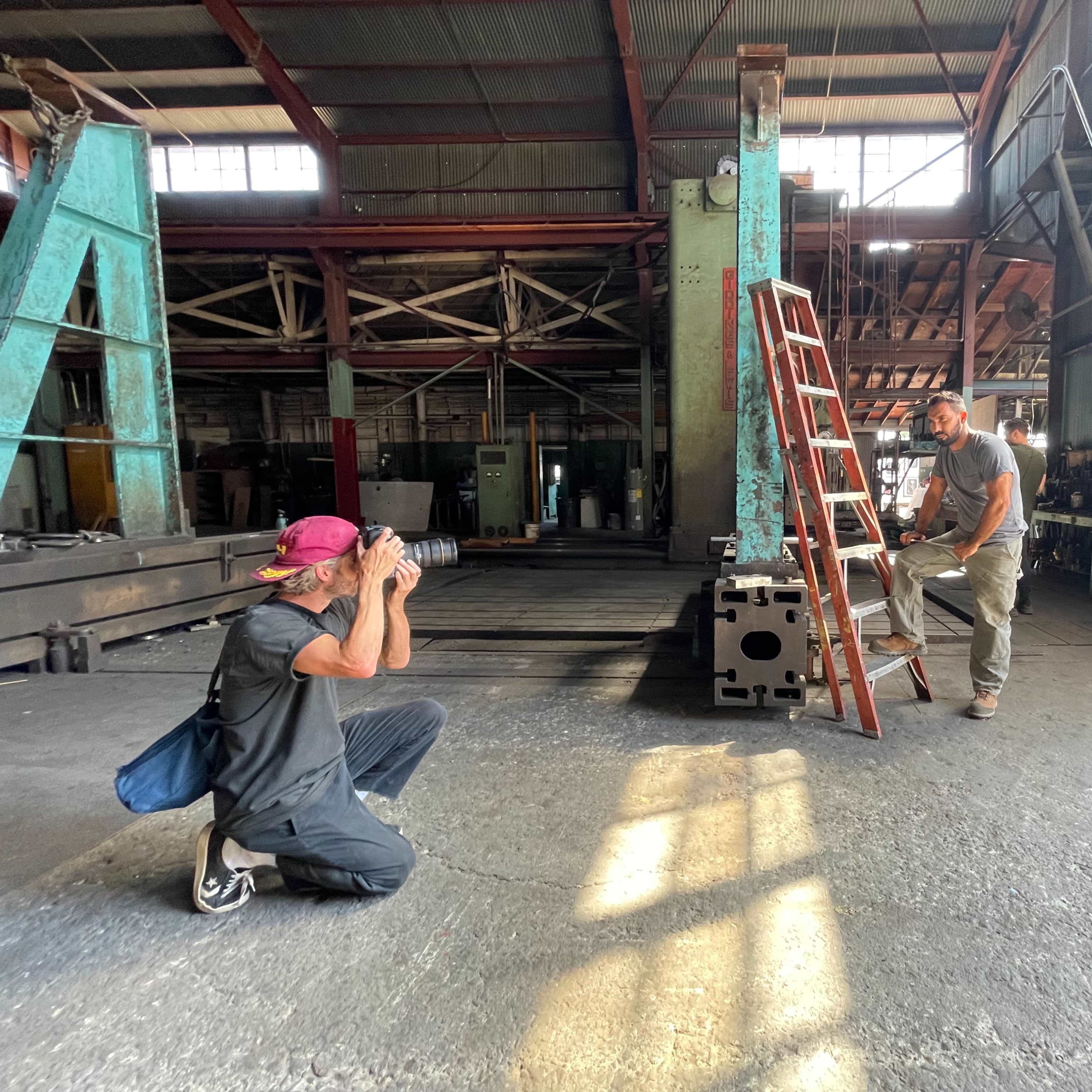 A person is crouching and taking a photograph of another person who is standing on a ladder in a spacious industrial setting.