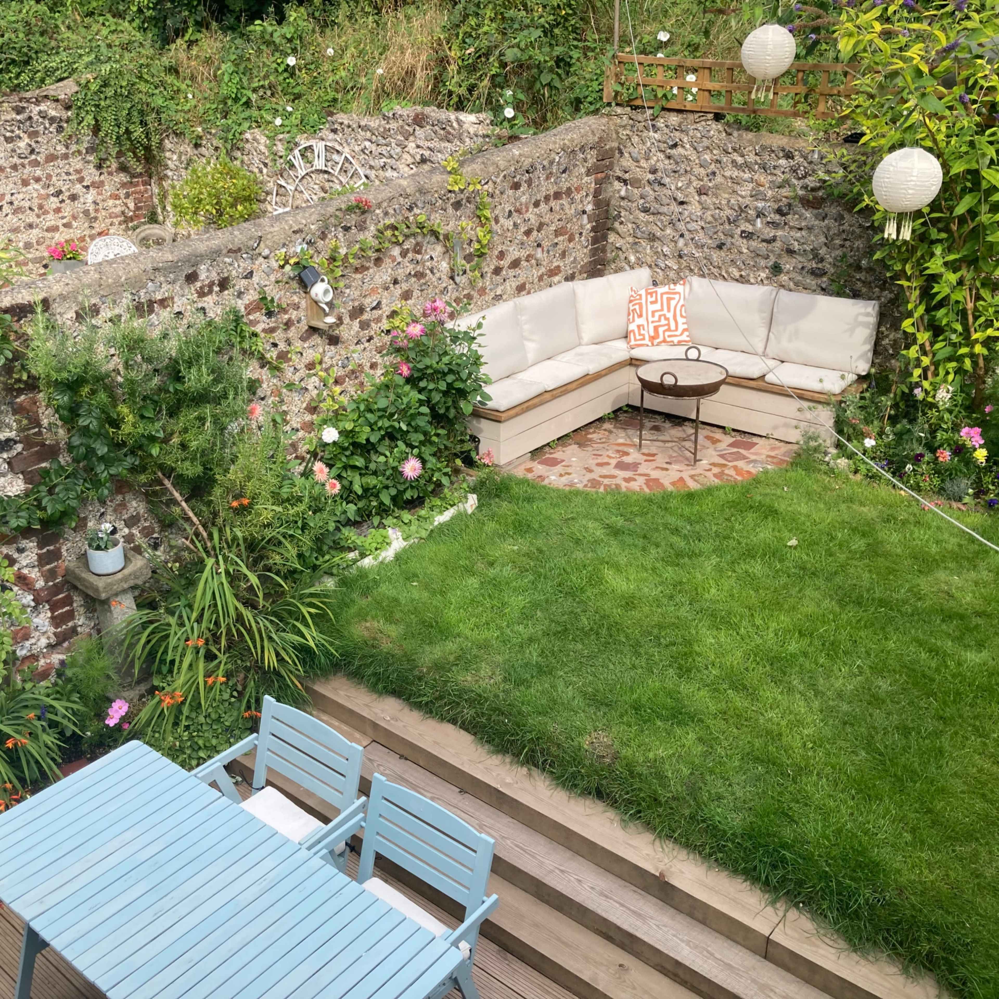 A small garden with a grassy area, a circular stone table, a corner seating arrangement, and a dining set on a wooden deck, surrounded by stone walls and plants.